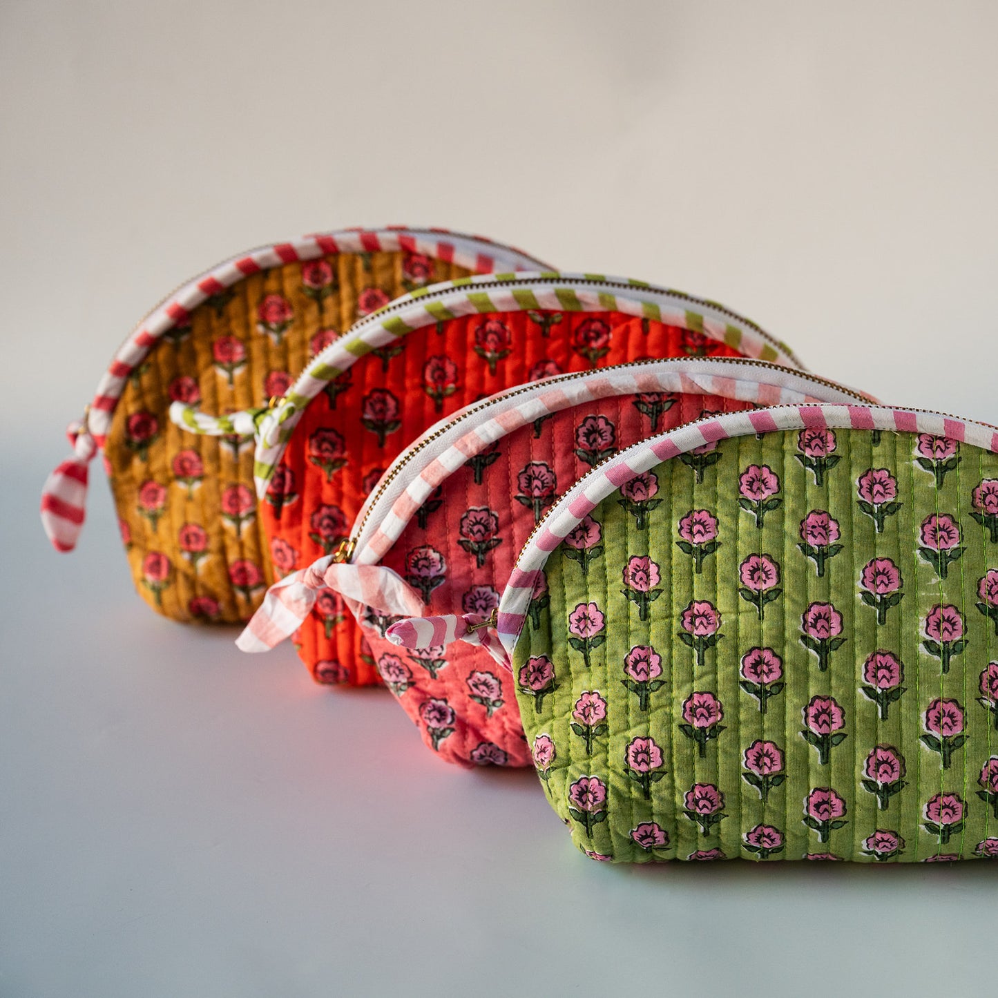 Three quilted zippered pouches in floral fabric, arranged diagonally on a light gray background. The pouches are mustard yellow with red flowers, coral red with pink flowers, and light green with darker green and pink flowers.