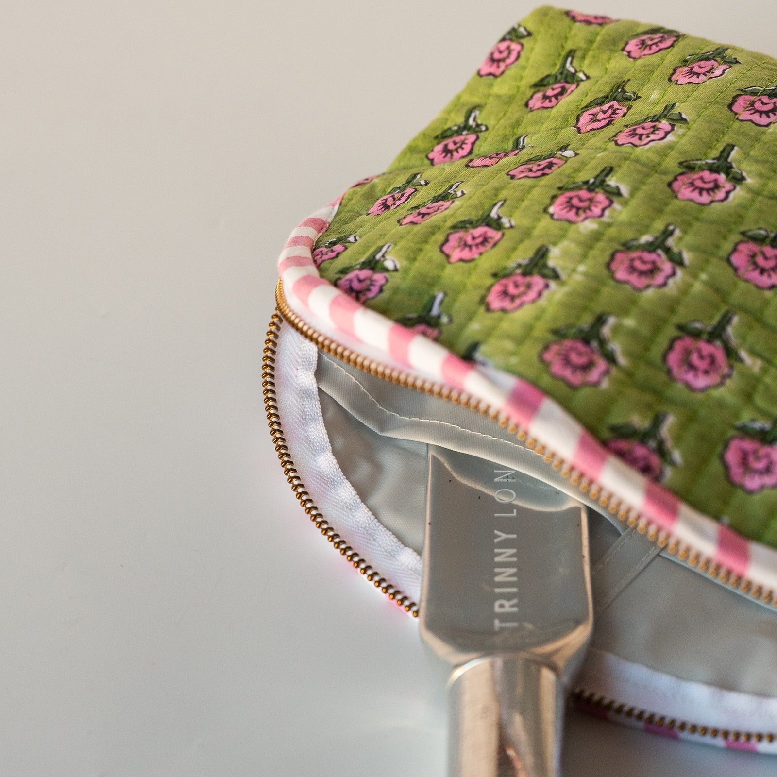 A close-up of a quilted fabric pouch with a green floral print, pink and white gingham trim, and a gold zipper, shown with a metal spoon resting inside.
