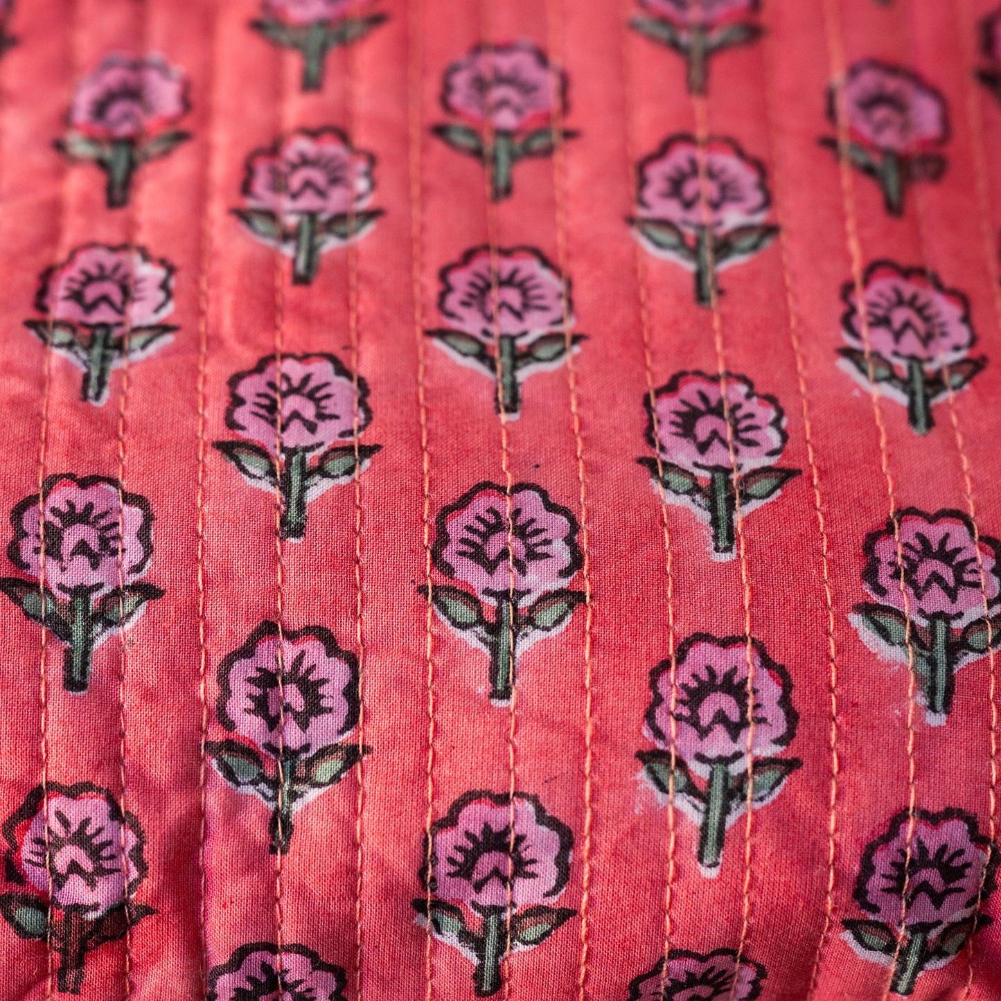 A close-up of a coral pink fabric with a repeating pattern of pink flowers and green leaves, featuring vertical hand-stitched seams.