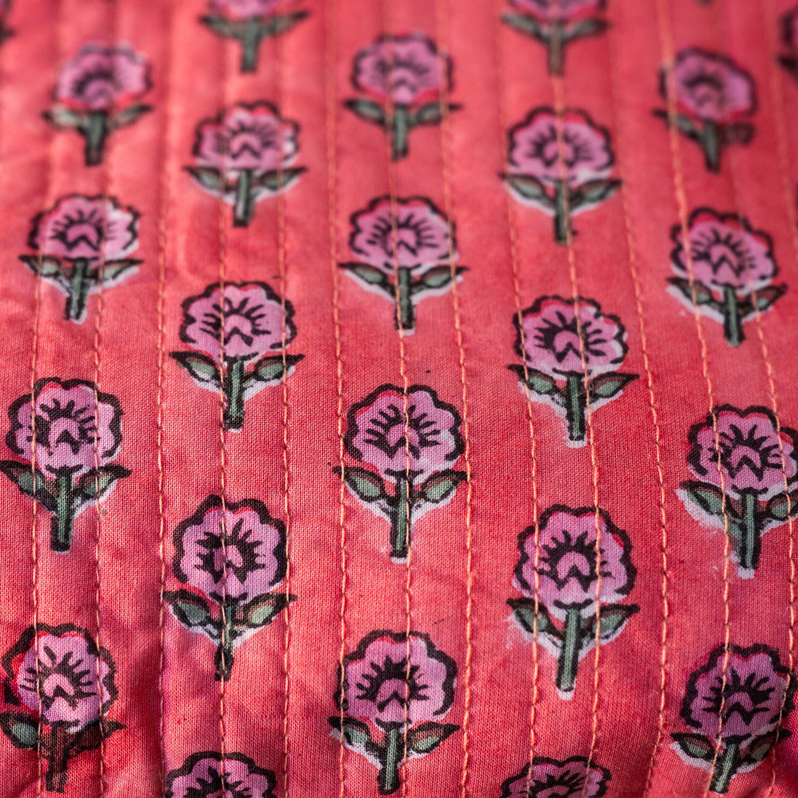A close-up of a coral pink fabric with a repeating pattern of pink flowers and green leaves, featuring vertical hand-stitched seams.