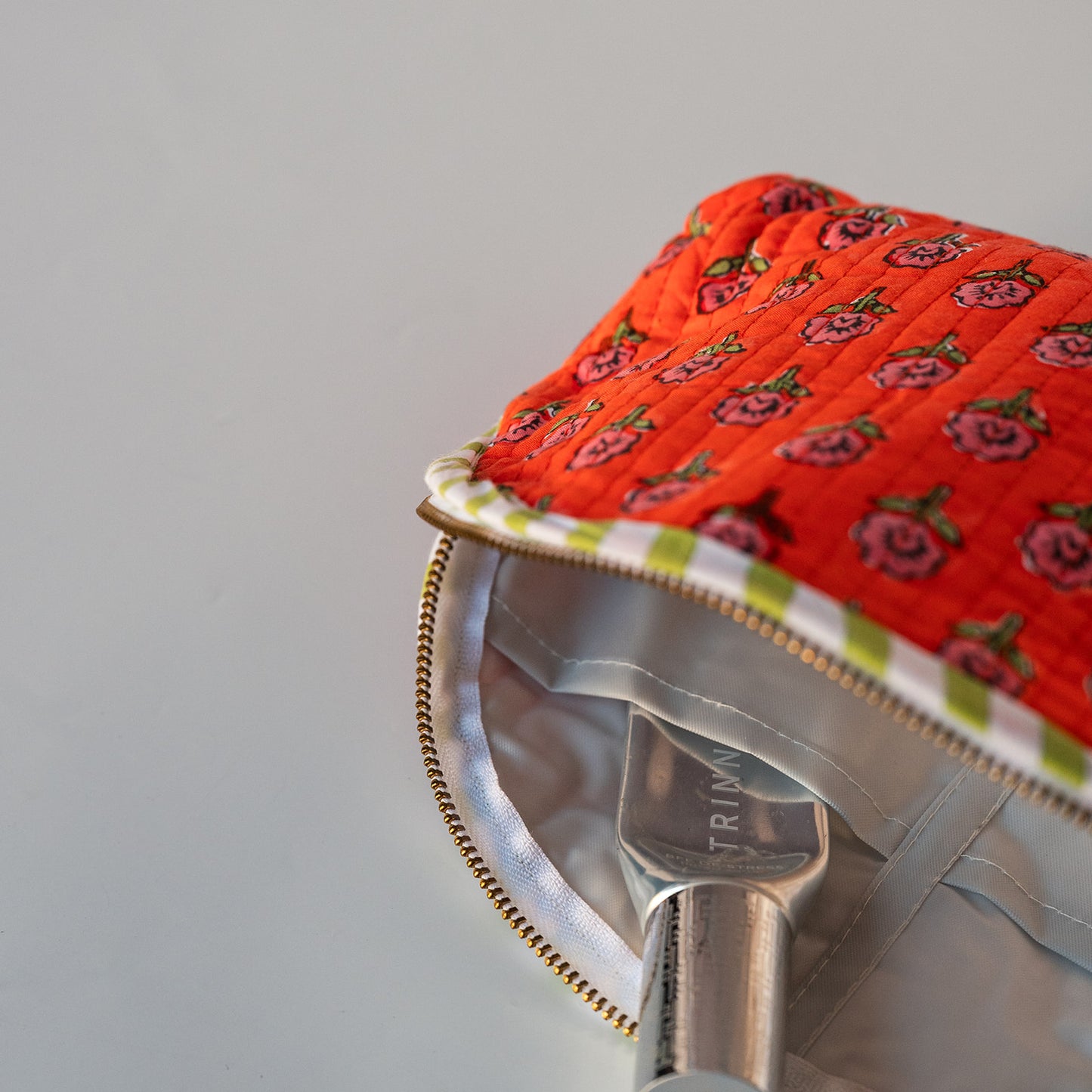 Close-up of a quilted red-orange fabric accessory with a pink rose pattern, gold zipper, and white gingham trim, shown with a silver metal handle inside on a light gray background.