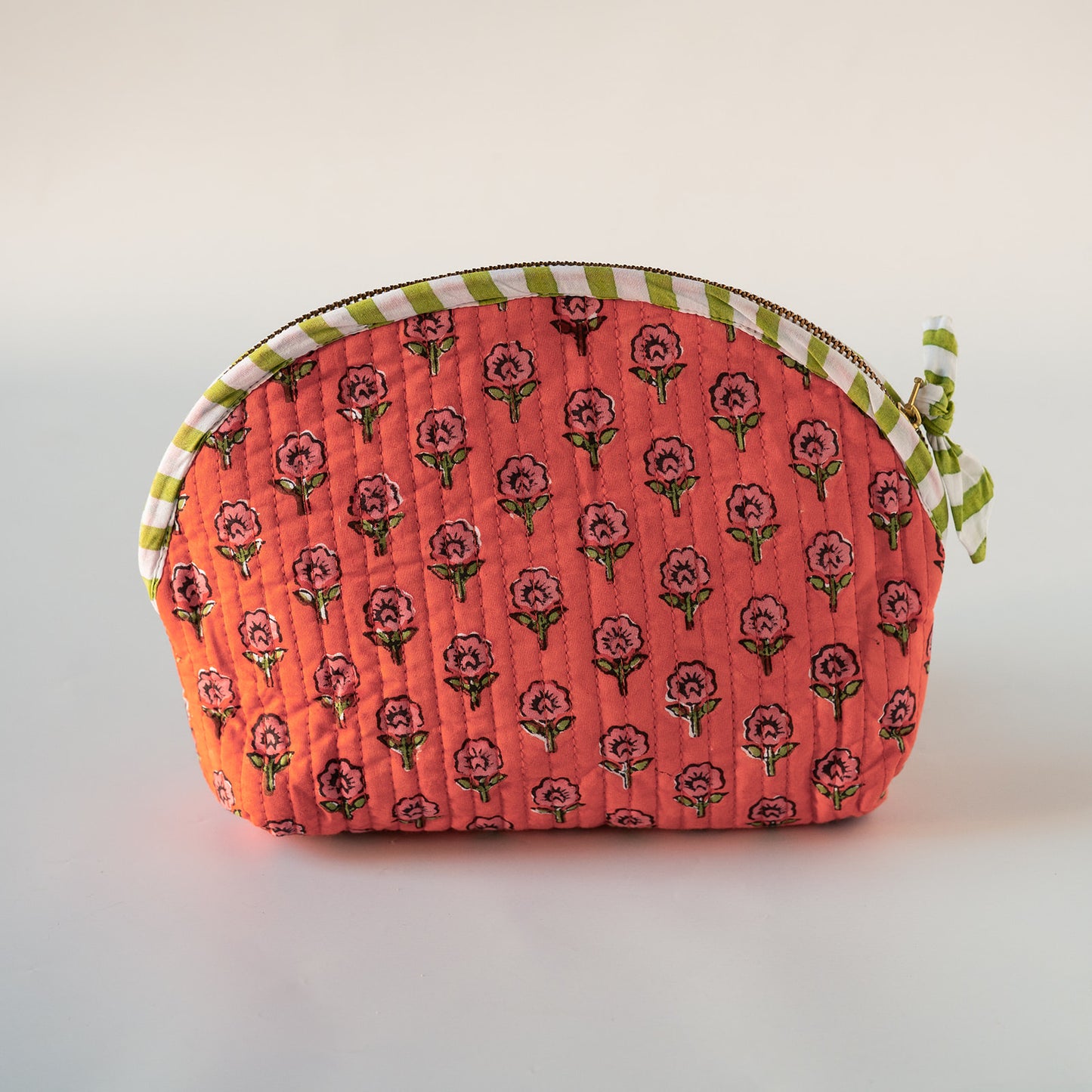 A quilted fabric cosmetic pouch with a coral pink background, a repeating pink rose pattern, and a green and white striped trim with a matching bow on the zipper pull, shown on a neutral light gray background.