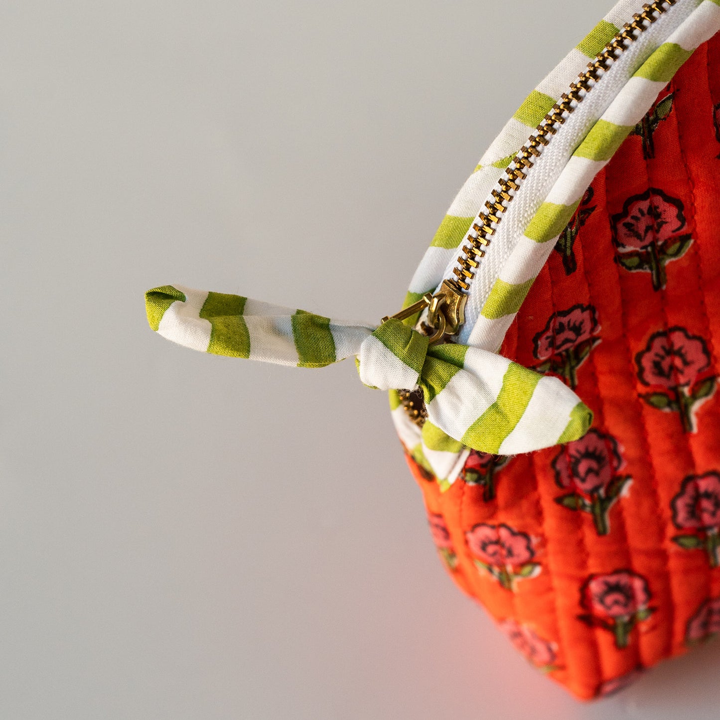 Close-up of a quilted fabric zippered pouch with a bright orange-red floral body, green-and-white striped zipper trim, and a matching striped fabric tie closure.