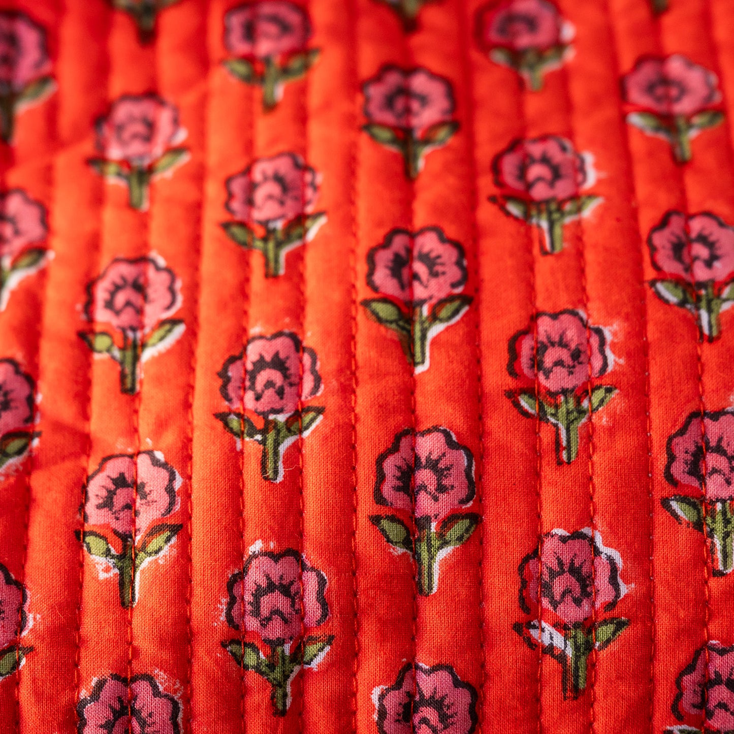 A close-up of a quilted fabric featuring a repeating pattern of pink flowers with green stems on a bright orange-red background.