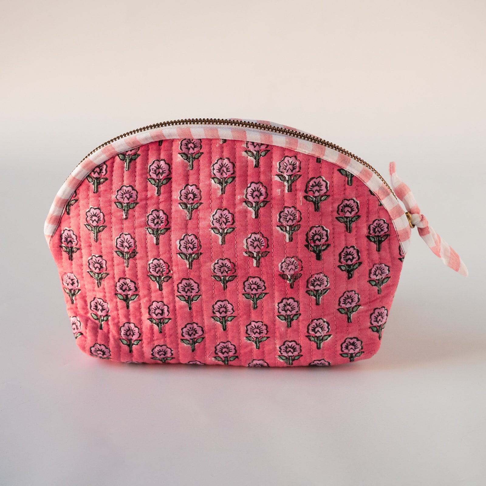 A quilted fabric cosmetic pouch with a pink background, featuring a repeating floral block print in dark green and purple, finished with a white and pink gingham trim and a gold zipper.