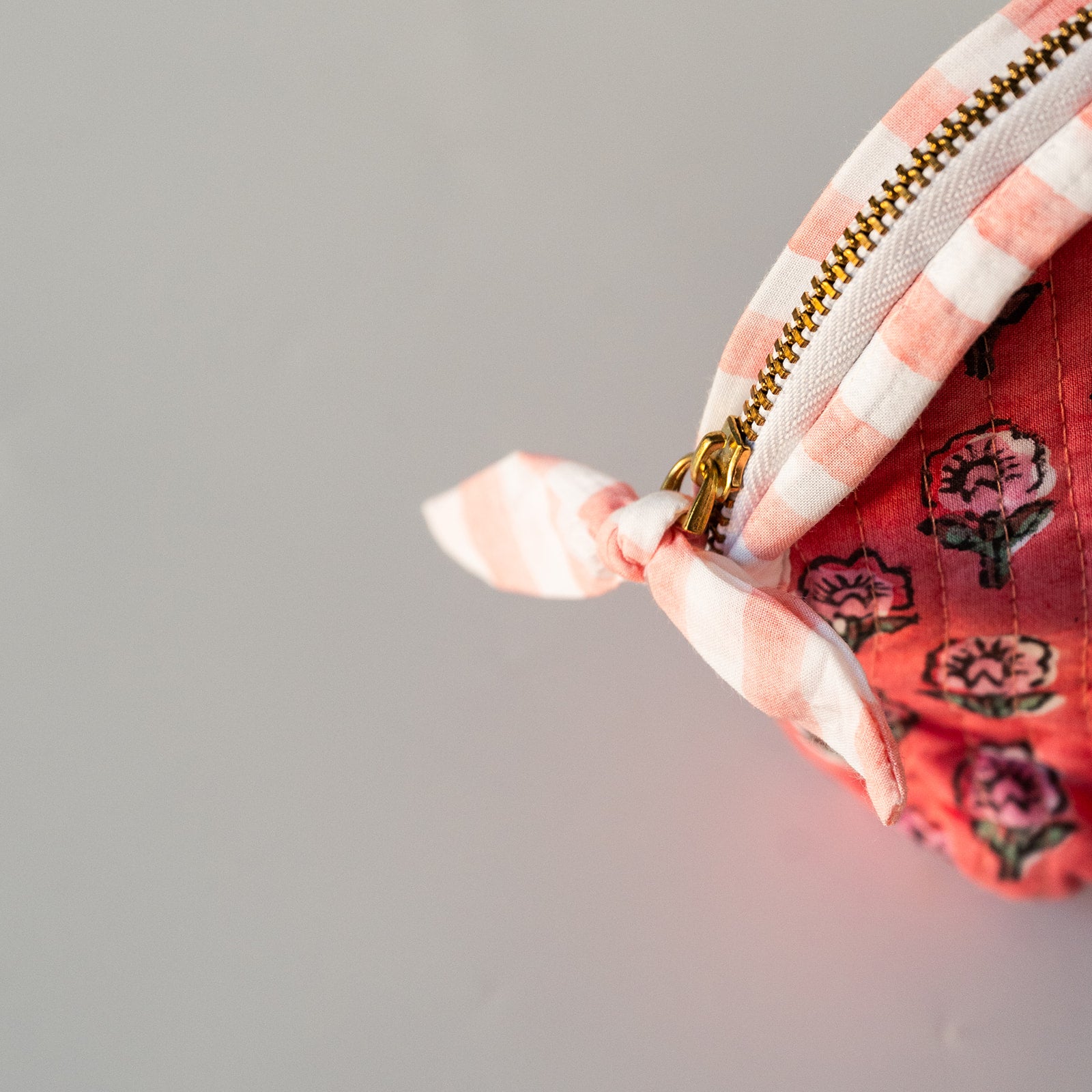 Close-up of a fabric zippered pouch corner showing a pink and white gingham zipper trim, a gold-tone zipper pull, and a tied pink-and-white fabric loop on a red floral quilted fabric body, set against a plain light gray background.