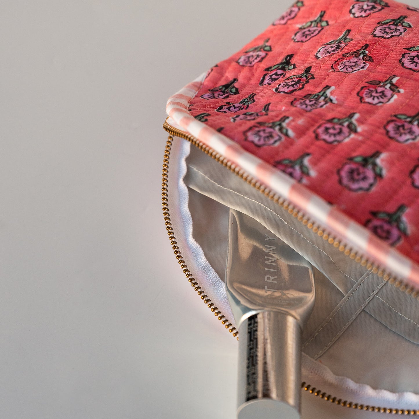 Close-up of a quilted cosmetic pouch with a pink floral pattern, gold zipper, and white lining, shown with a silver-handled applicator.