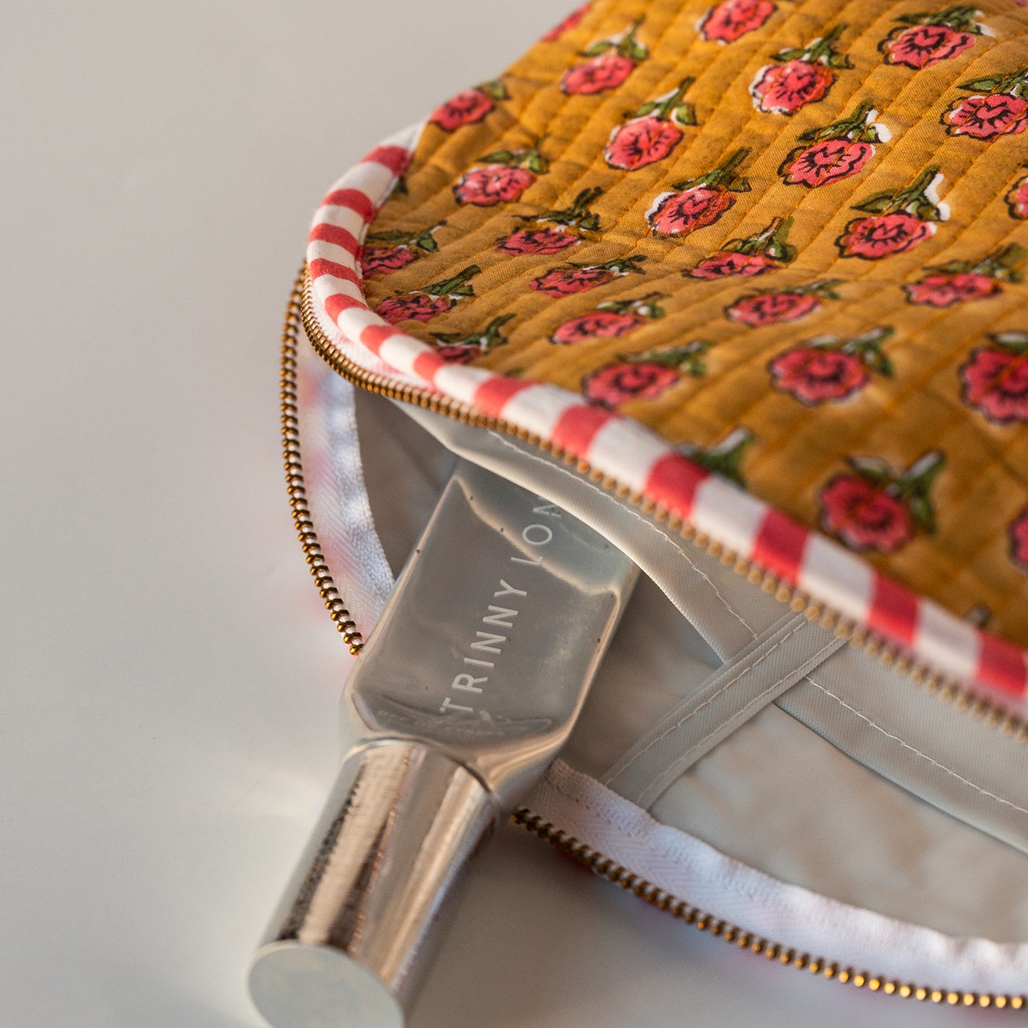 A close-up of a quilted cosmetic pouch in mustard yellow with a pink rose pattern, featuring a red and white striped zipper trim and a gold-tone zipper. A silver metallic bottle labeled 'TRINNY WOOD' is partially inserted into the pouch.