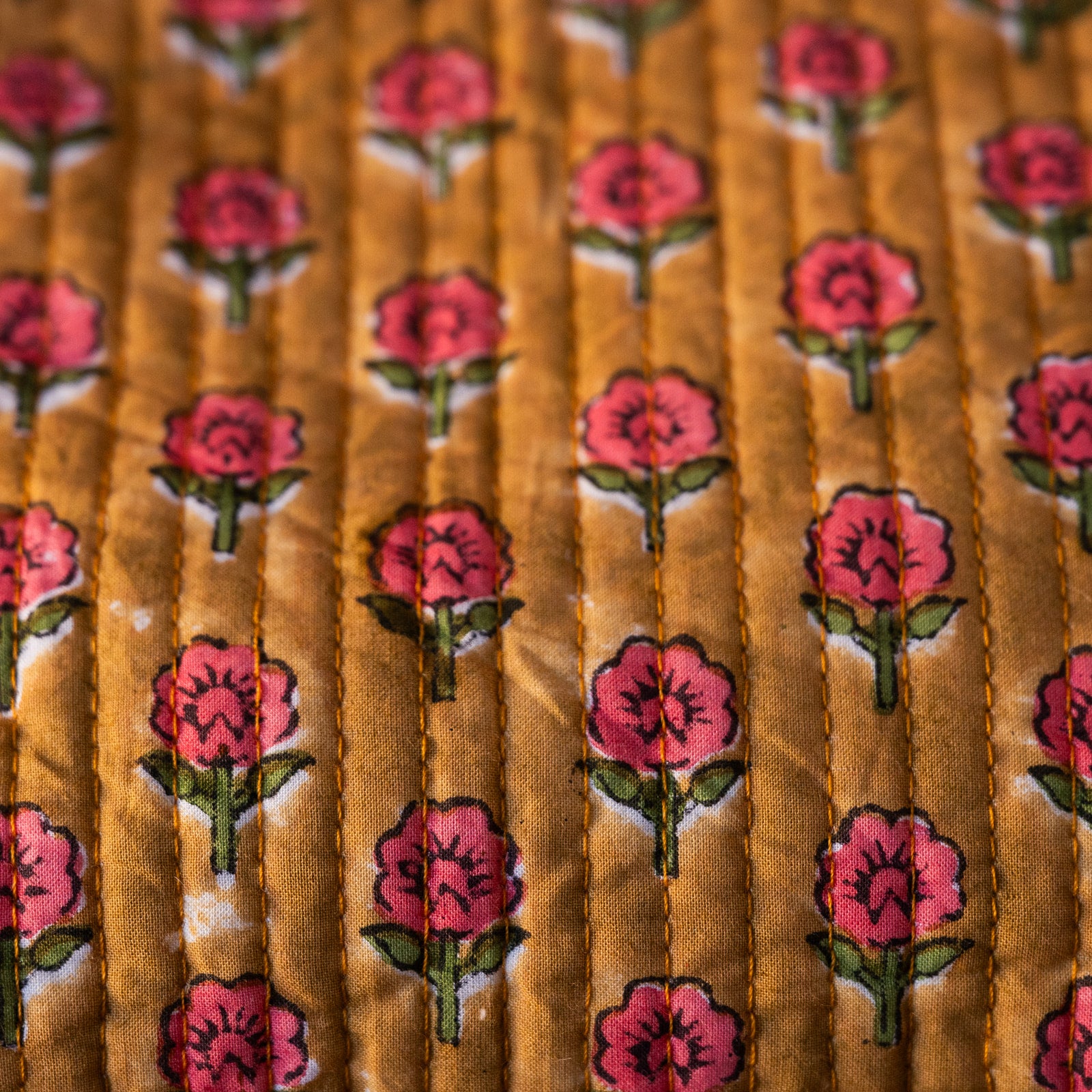 A close-up of a quilted fabric with a repeating pattern of pink flowers with green stems on a mustard yellow background.