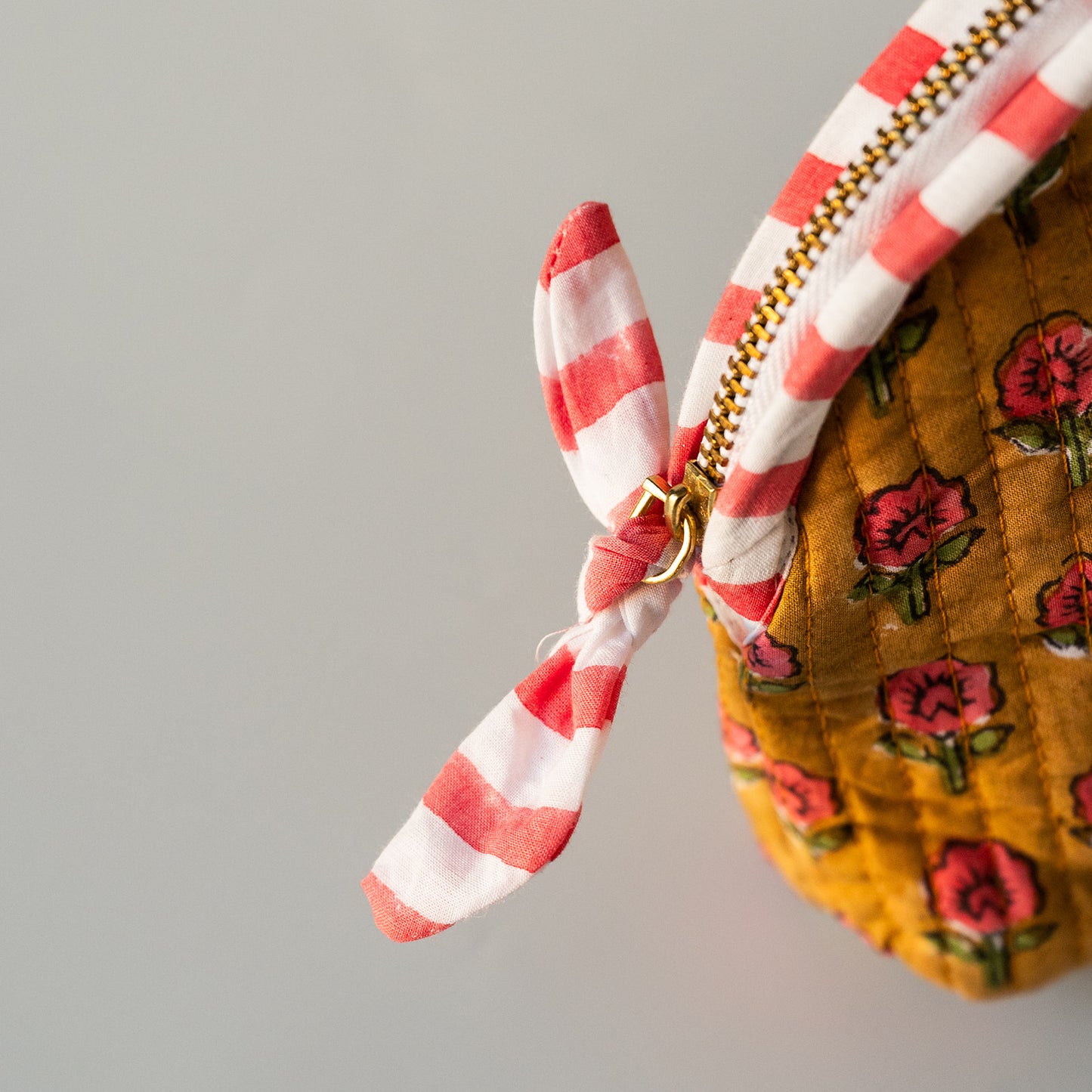 Close-up of a quilted mustard yellow pouch with a red and white striped fabric zipper pull tied in a bow, set against a plain light gray background.