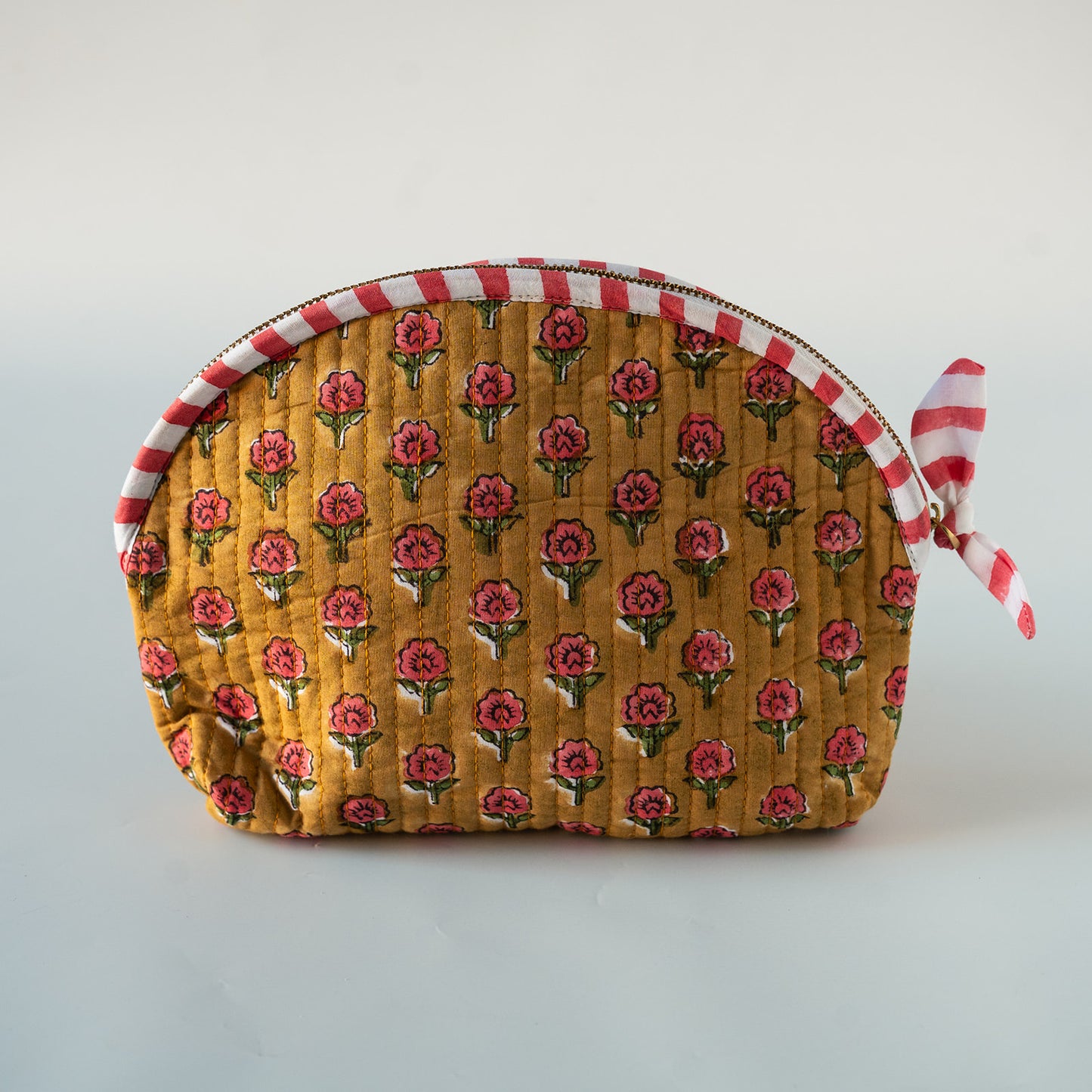A quilted fabric cosmetic pouch with a mustard yellow background, featuring a repeating red and green floral block print. The pouch has a red and white striped zipper trim and a matching red and white striped ribbon tie on the side, photographed on a neutral light gray background.
