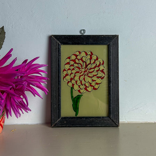 Framed artwork of a red and yellow flower on a light blue wall with a pink flower in the foreground.