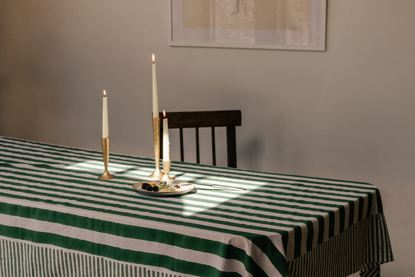 A green and white striped tablecloth draped over a dining table, styled with gold candlesticks and a small plate, with a dark wooden chair and a neutral wall in the background.