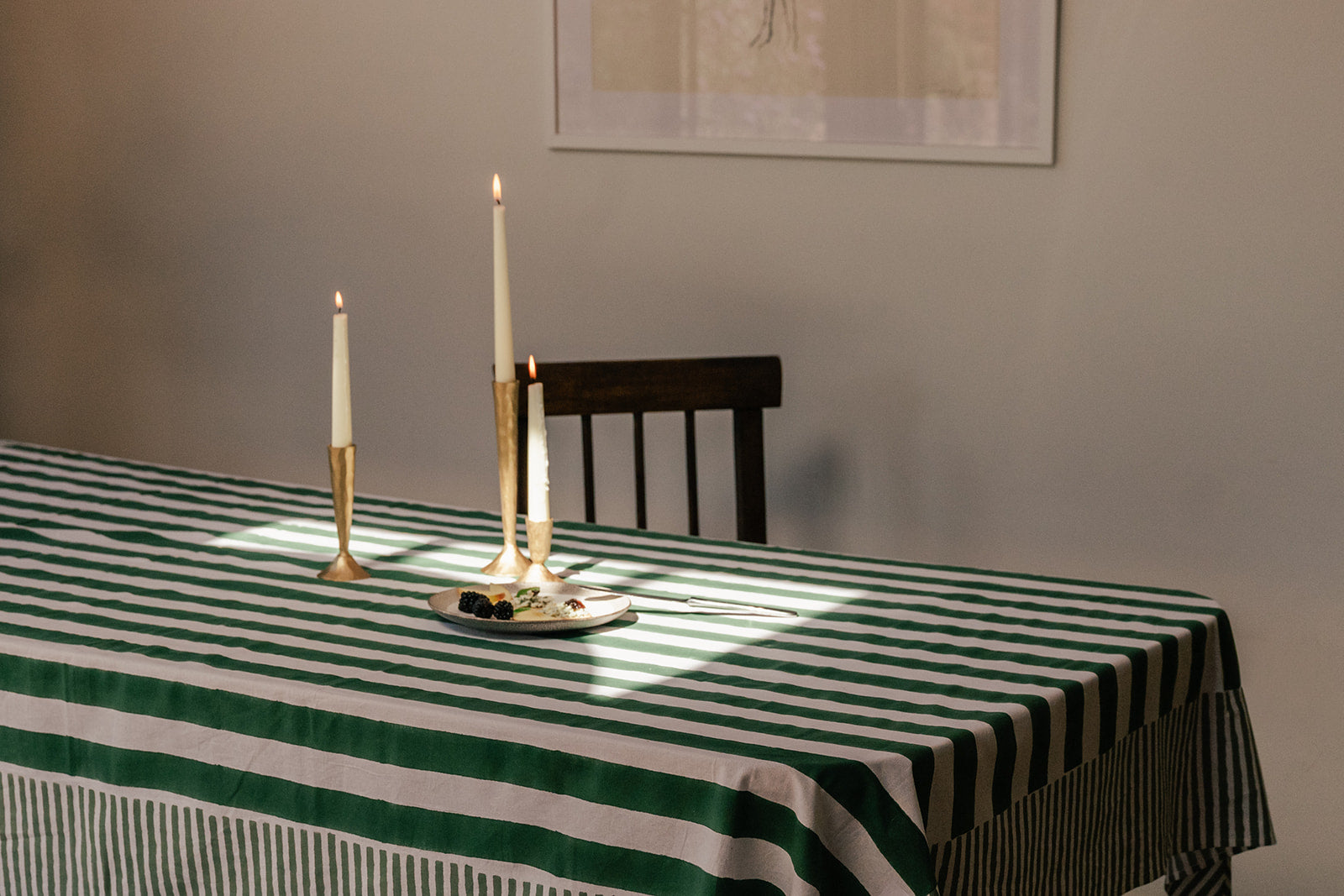 A green and white striped tablecloth draped over a dining table, styled with gold candlesticks and a small plate, with a dark wooden chair and a neutral wall in the background.