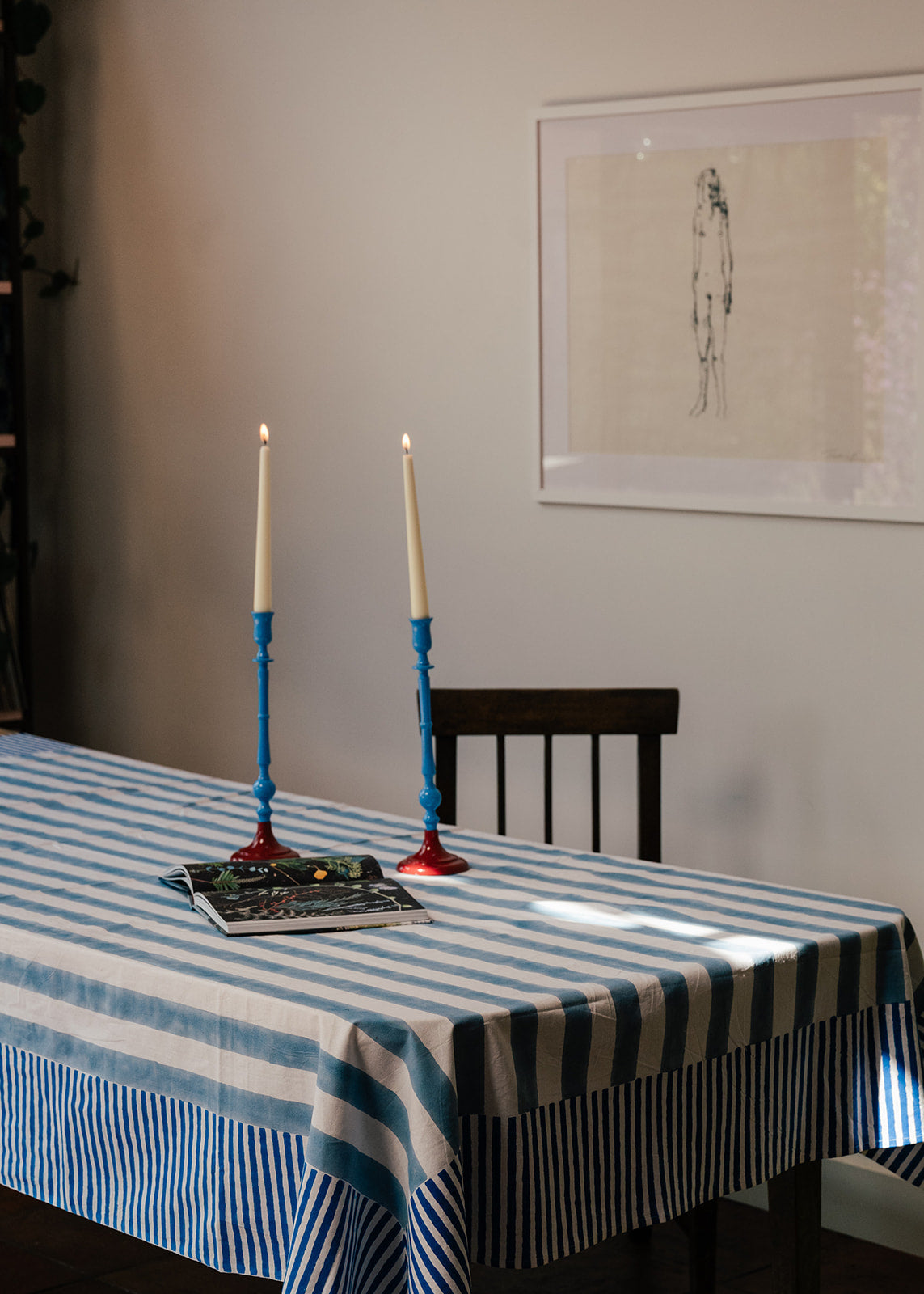 A blue and white striped tablecloth draped over a dining table, styled with two blue candlesticks holding lit taper candles and a magazine. A dark wooden chair is behind the table, and a framed abstract print hangs on the beige wall.