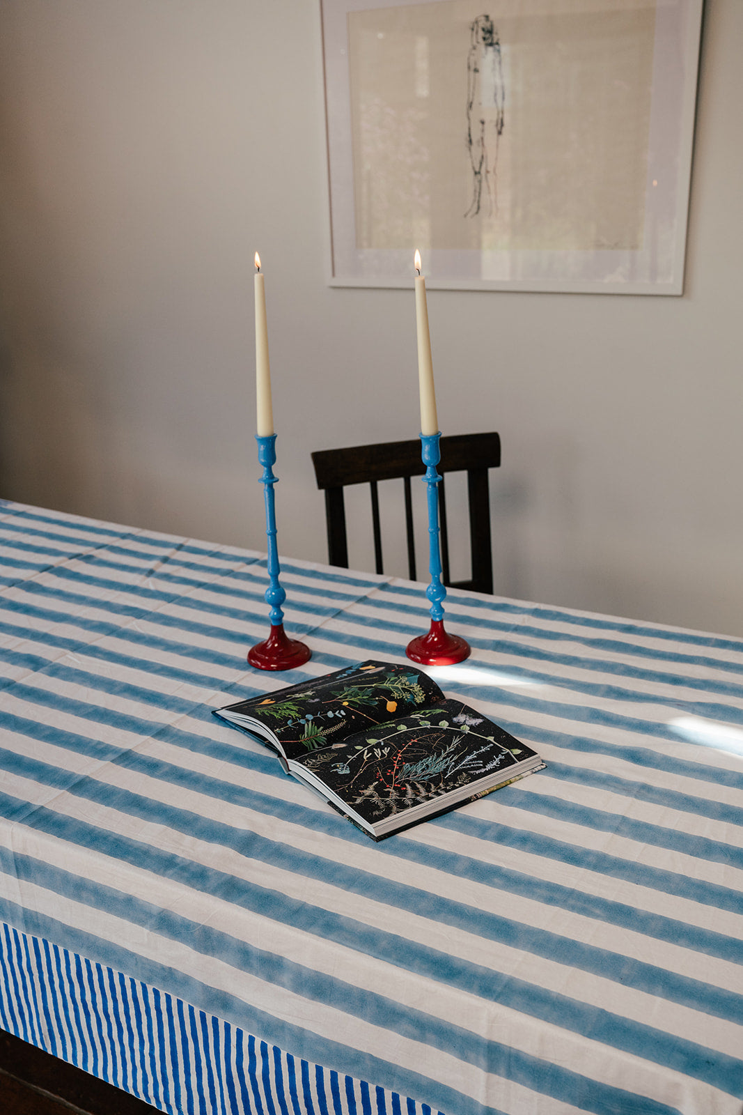 A blue and white striped tablecloth on a dining table, with two blue candlesticks holding lit white taper candles and an open illustrated book in front. A dark wooden chair and framed wall art are visible in the background.