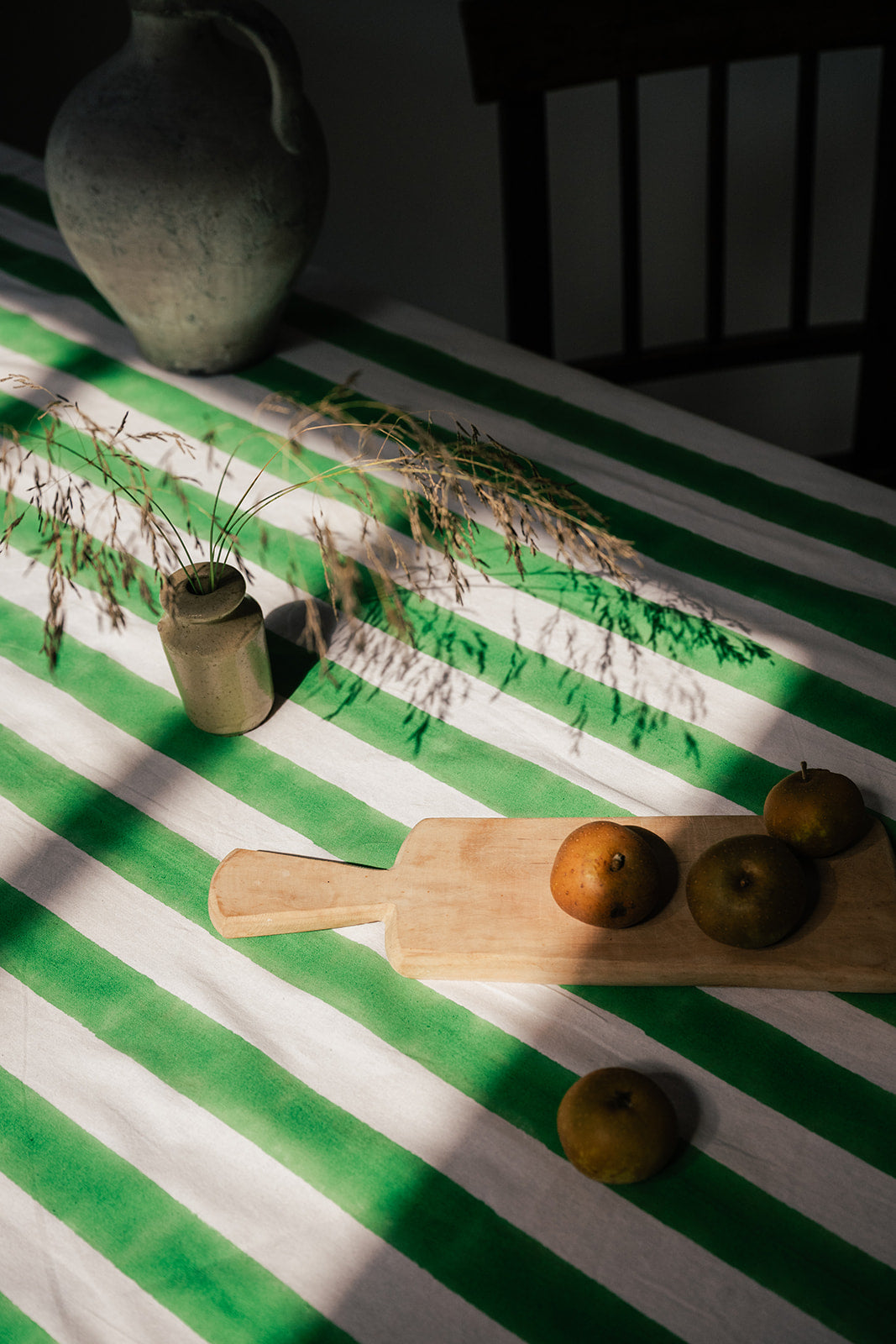 A green and white striped tablecloth draped over a table, styled with a wooden cutting board, three oranges, a small vase with dried grass, and a large ceramic jug in the background under dramatic, shadowed sunlight.