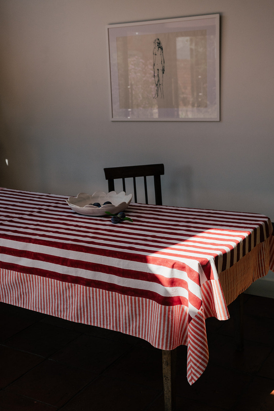 A striped tablecloth with alternating red and white horizontal bands draped over a table in a dining room setting, with a chair, bowl, and framed wall art visible.
