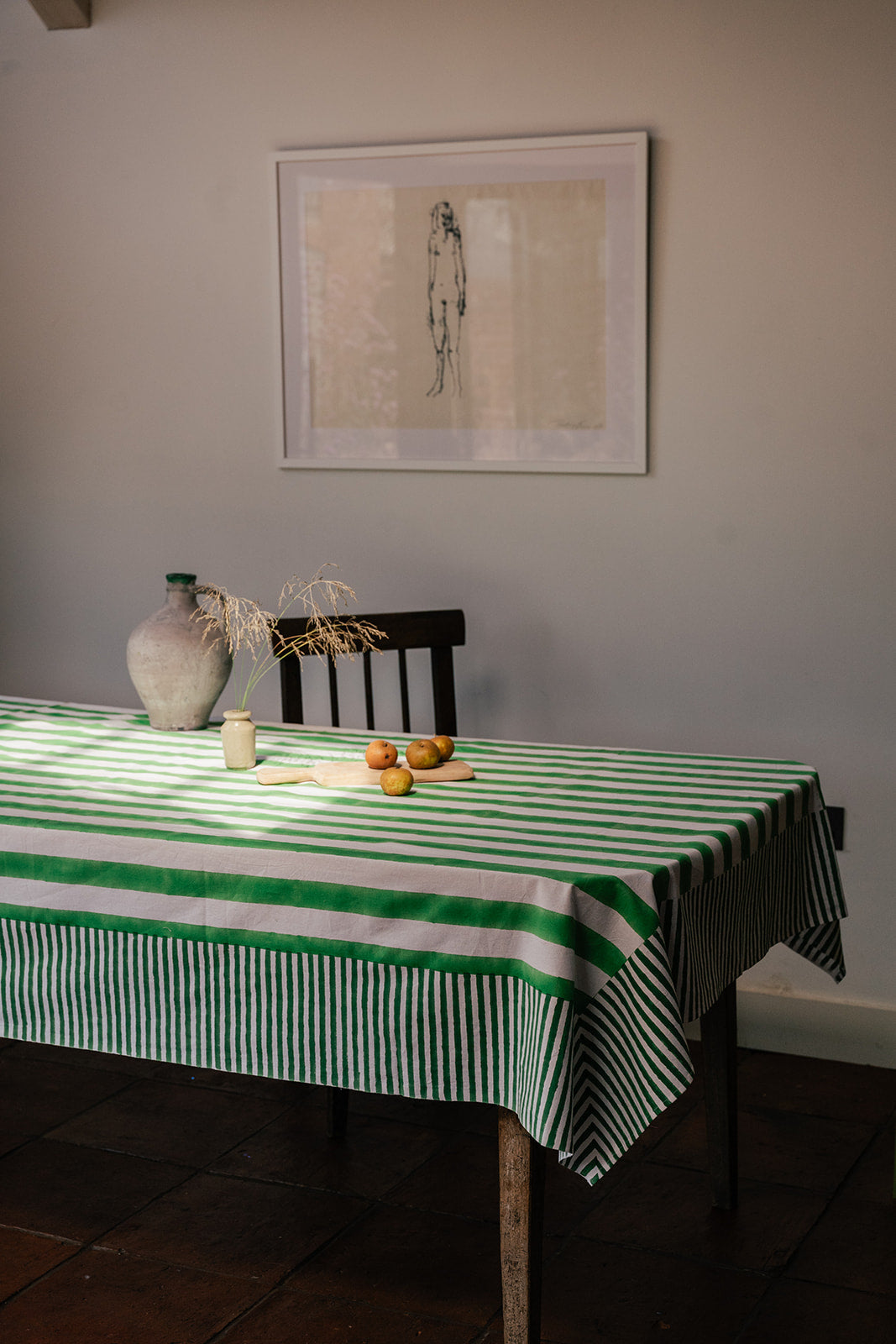 A green and white striped tablecloth draped over a wooden dining table in a styled room setting, with a vase, dried stems, and small fruit on the table, and a framed art print on the wall behind.