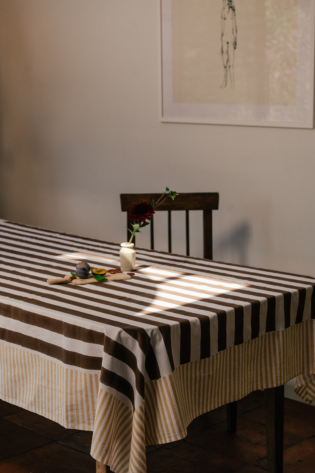 A rectangular tablecloth with alternating dark brown and cream horizontal stripes, draped over a wooden dining table in a warmly lit room. A small vase with a single red flower and a small wooden spoon sit on the table, with a dark wooden chair behind it.