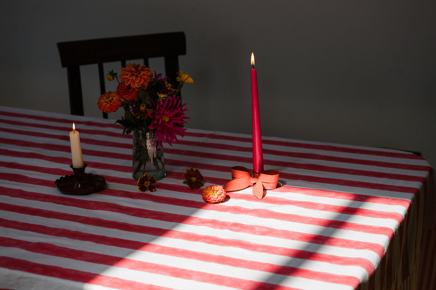 A red and white striped tablecloth on a table, styled with a glass vase of orange and pink flowers, a lit red taper candle in a decorative holder, and another lit white taper candle in a dark wooden chair in the background.