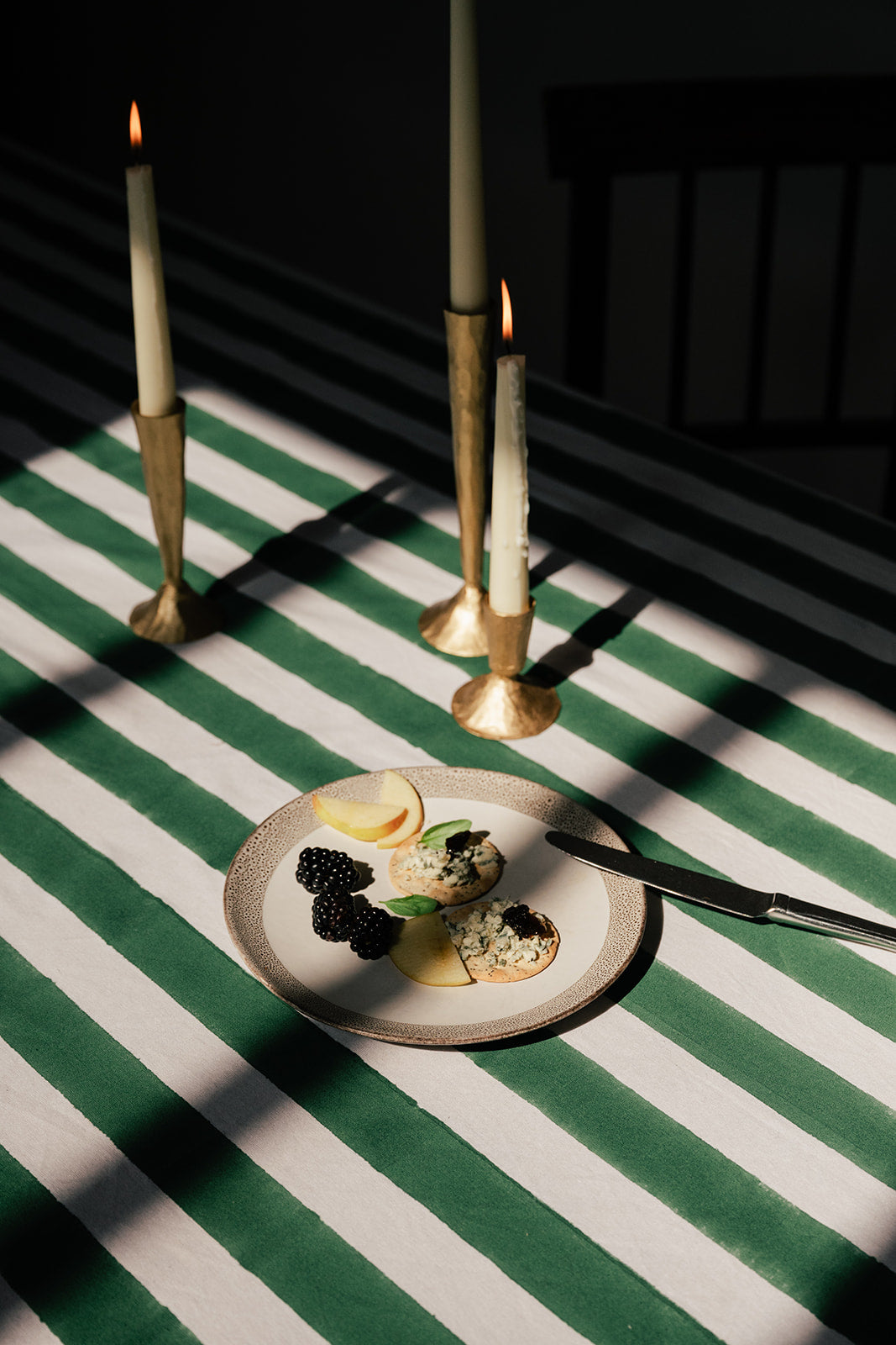 A styled tabletop scene with a small plate of food on a green-and-white striped tablecloth, alongside three lit taper candles in gold holders against a dark background.