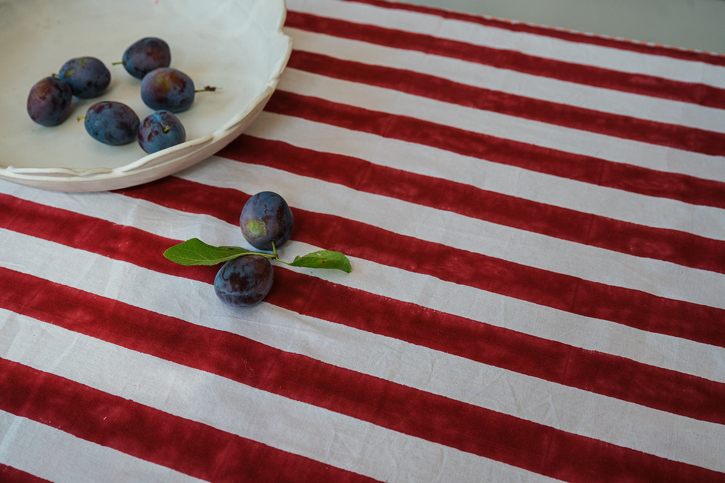 A red and white striped tablecloth with a velvet-like texture, styled with a white plate of plums and a small green leafy sprig on a light surface.