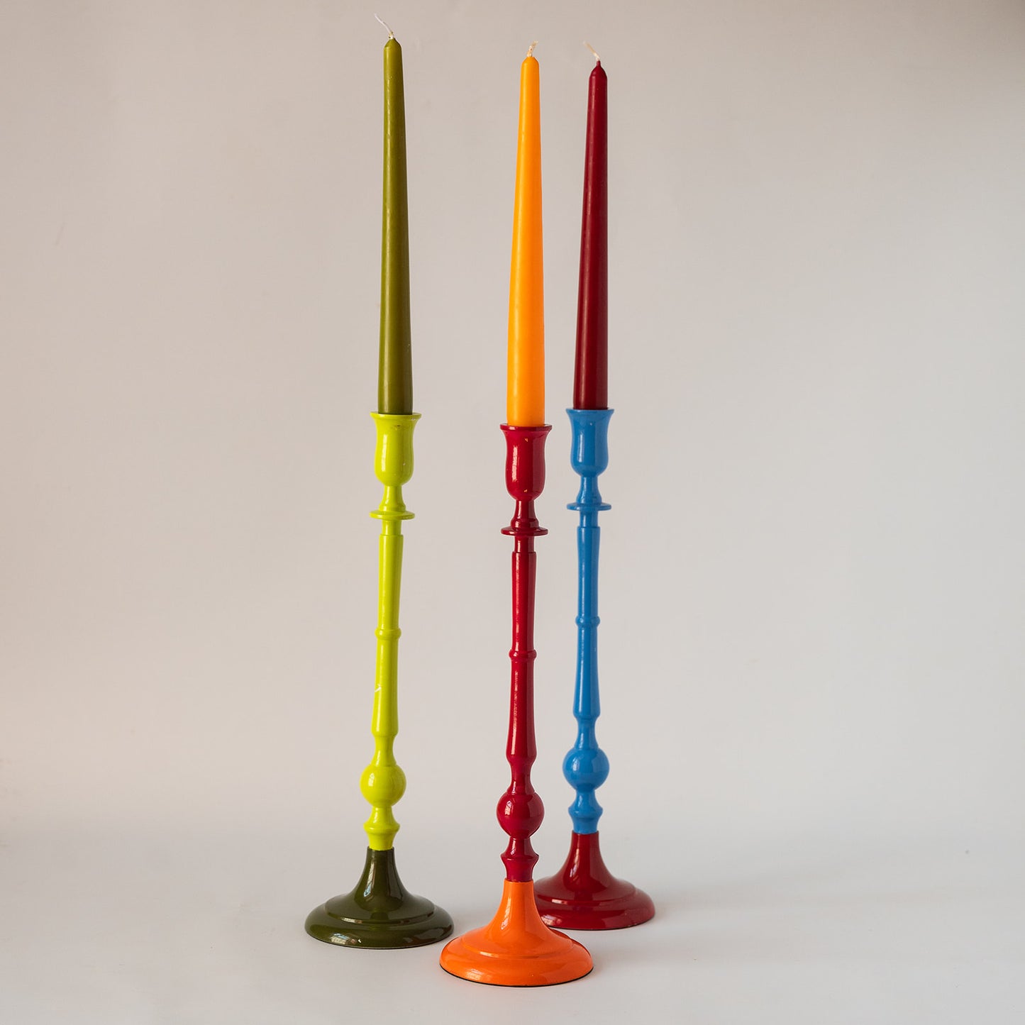Three tall taper candles in different colors displayed on matching painted candlesticks against a plain light gray background. The candlesticks are olive green, orange, and red, with the candles being dark olive green, bright orange, and dark red.