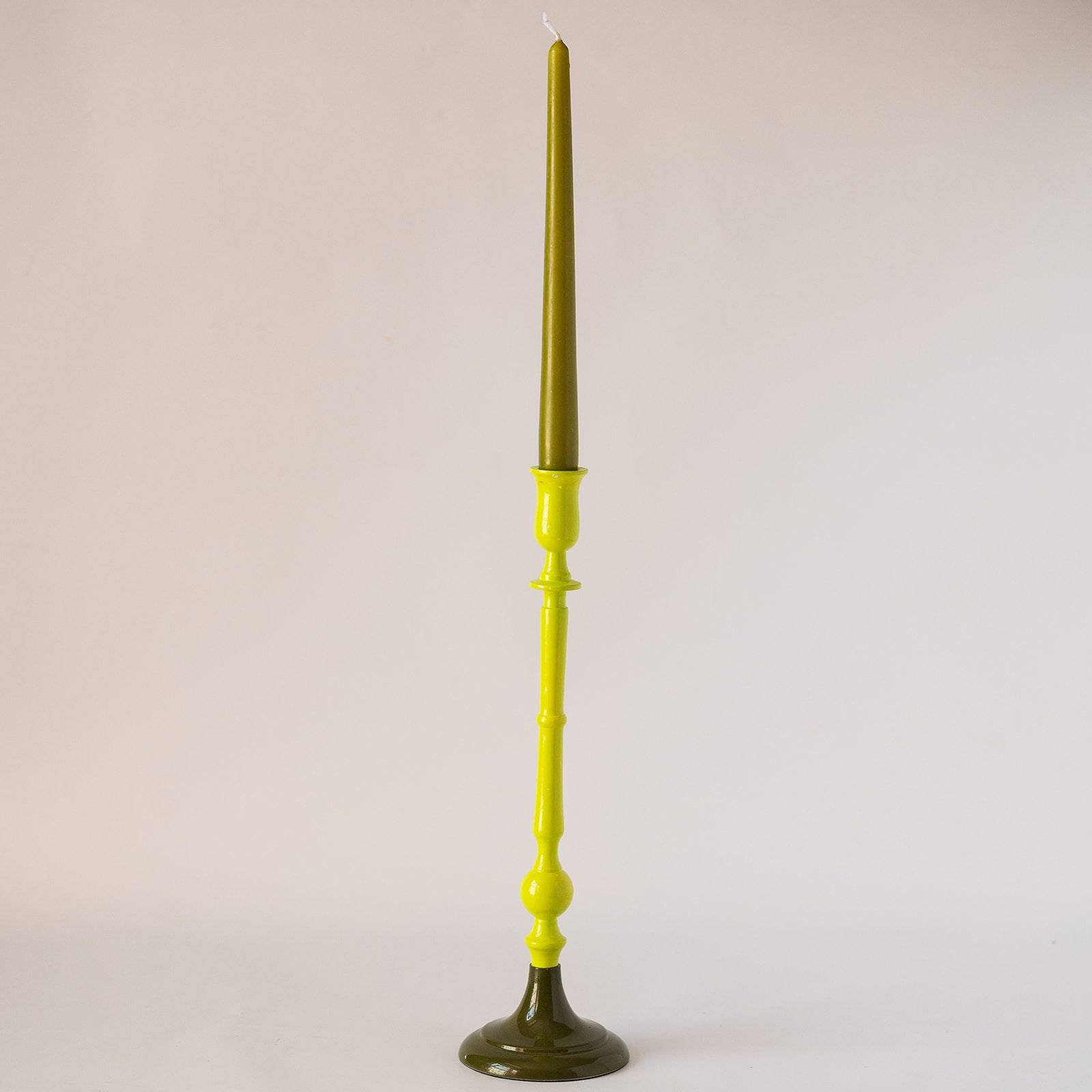 A tall, slender candlestick holder with a bright lime green stem and dark olive green base and top, holding a lit taper candle, set against a plain light gray background.
