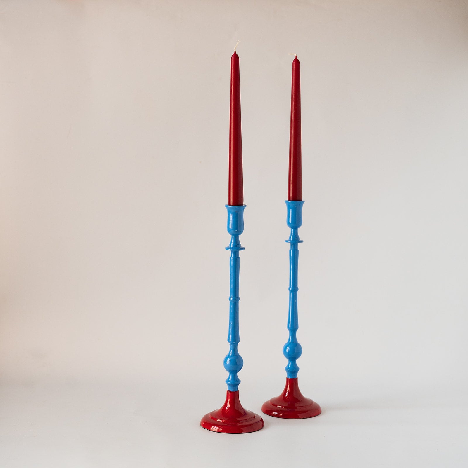 Two tall candlesticks with red tops and blue stems, standing on a light neutral background.