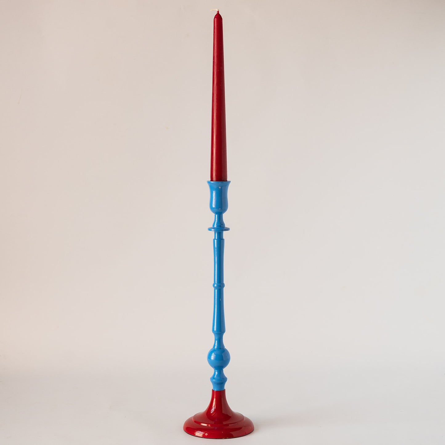 A tall candlestick holder with a glossy red base and taper, paired with a glossy blue stem, set against a plain light gray background.