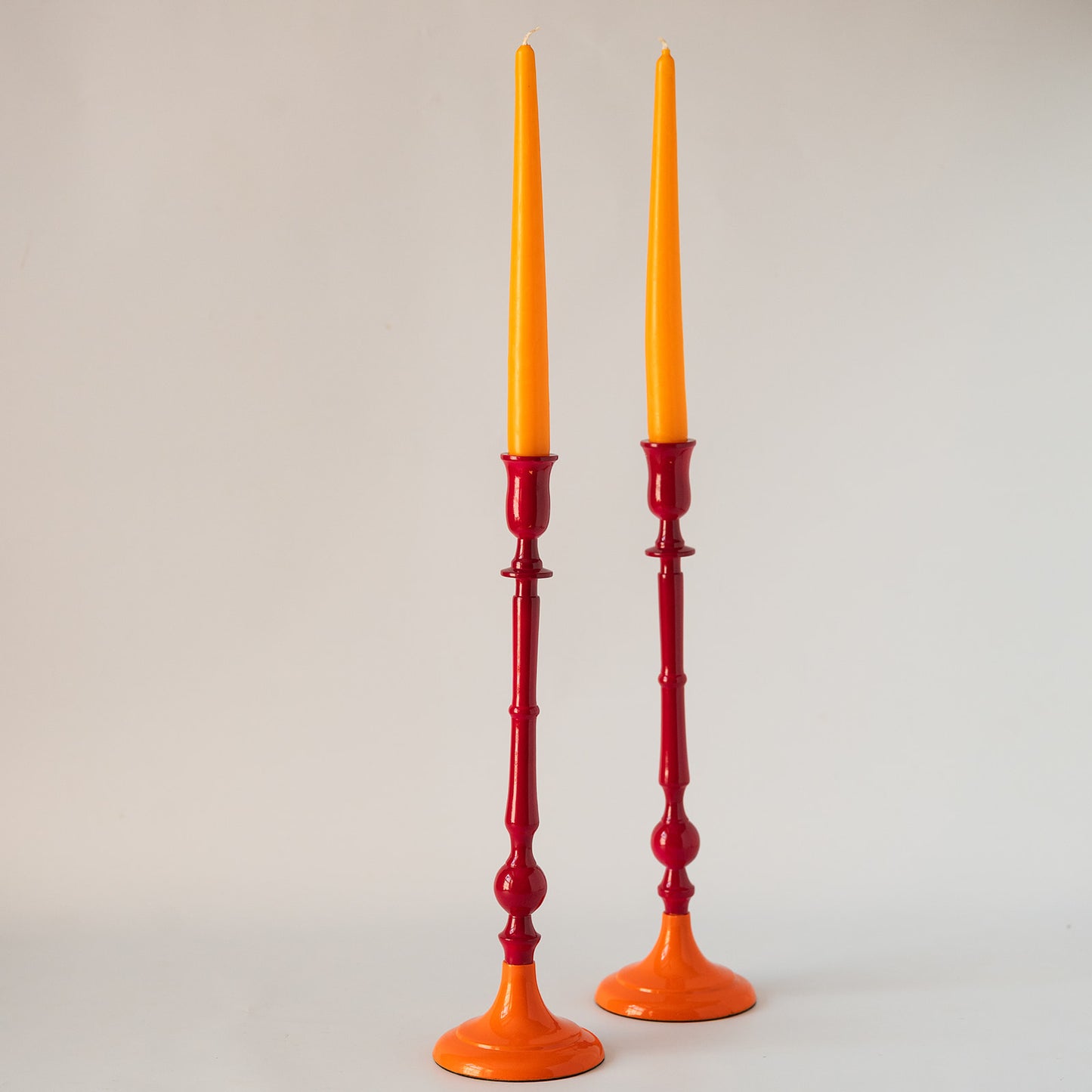 A pair of tall candlestick holders with bright orange bases and taper candles inserted, photographed against a plain light gray studio background.
