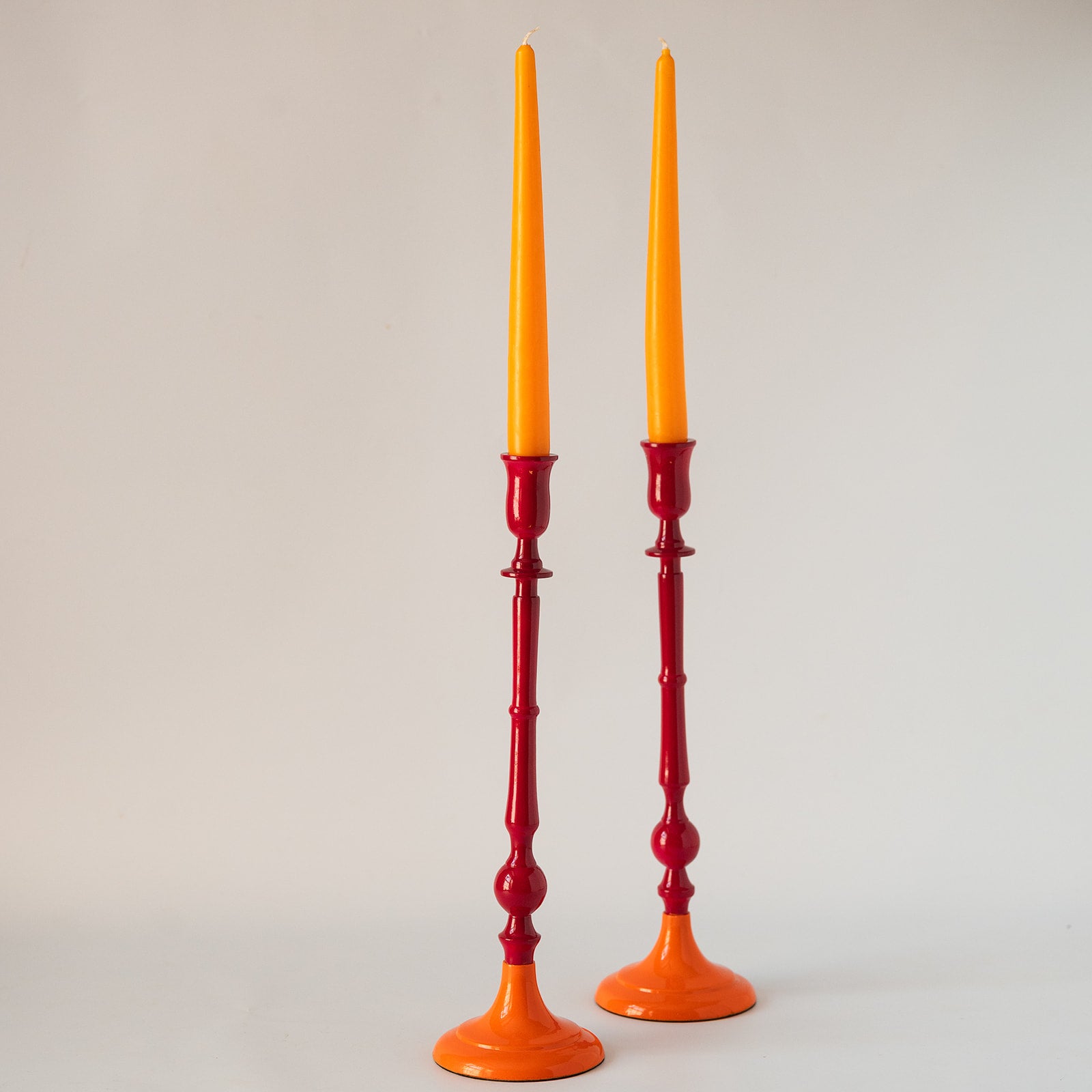 A pair of tall candlestick holders with bright orange bases and taper candles inserted, photographed against a plain light gray studio background.
