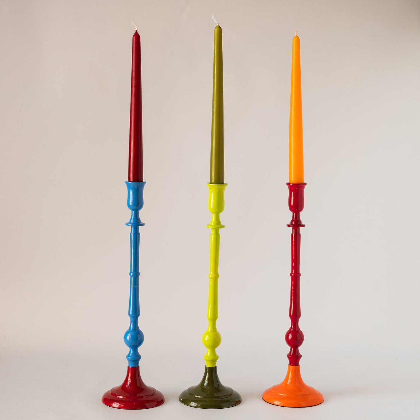 Three tall decorative candlesticks standing upright against a plain light background. The left candlestick has a red taper candle and a blue-and-red base.