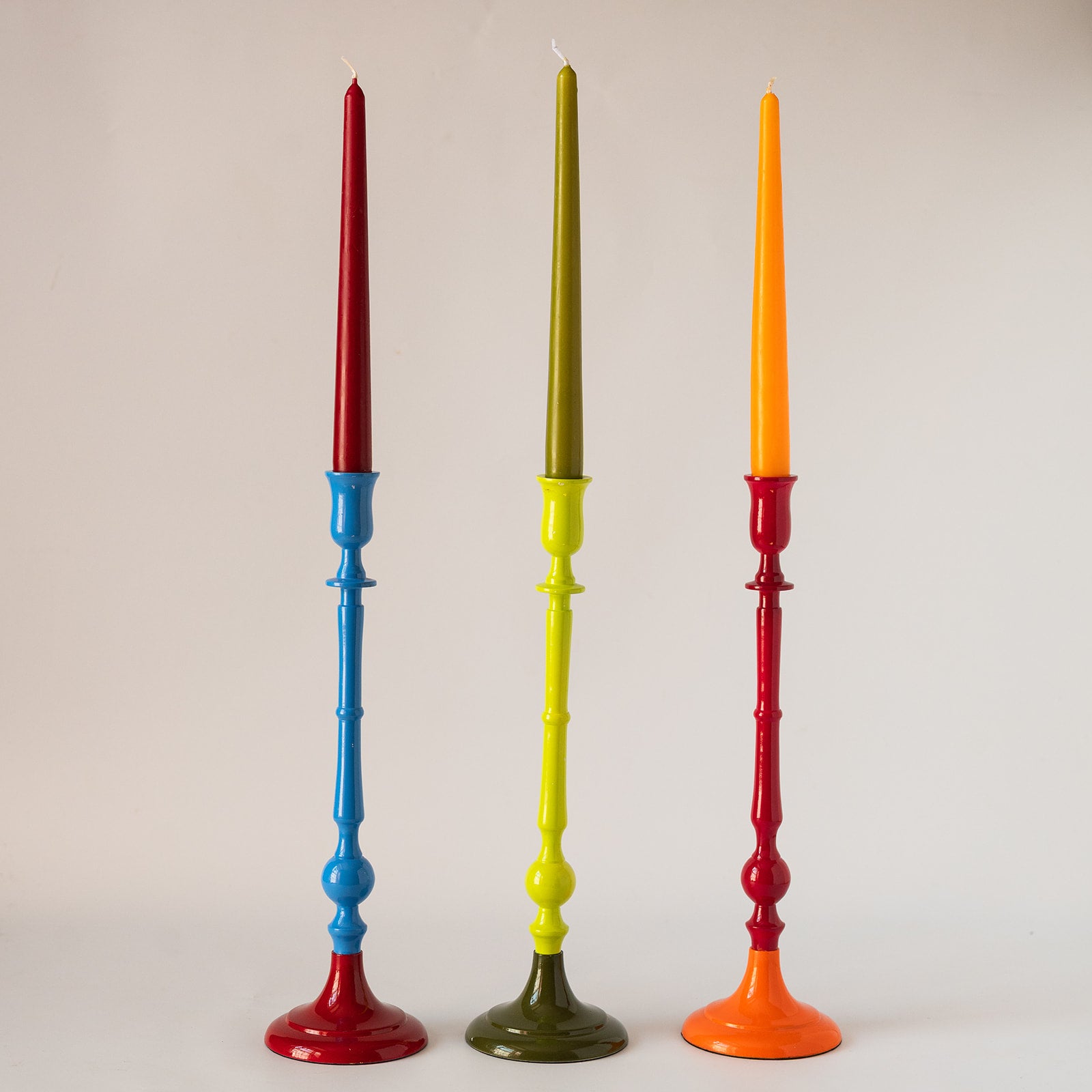 Three tall decorative candlesticks standing upright against a plain light background. The left candlestick has a red taper candle and a blue-and-red base.