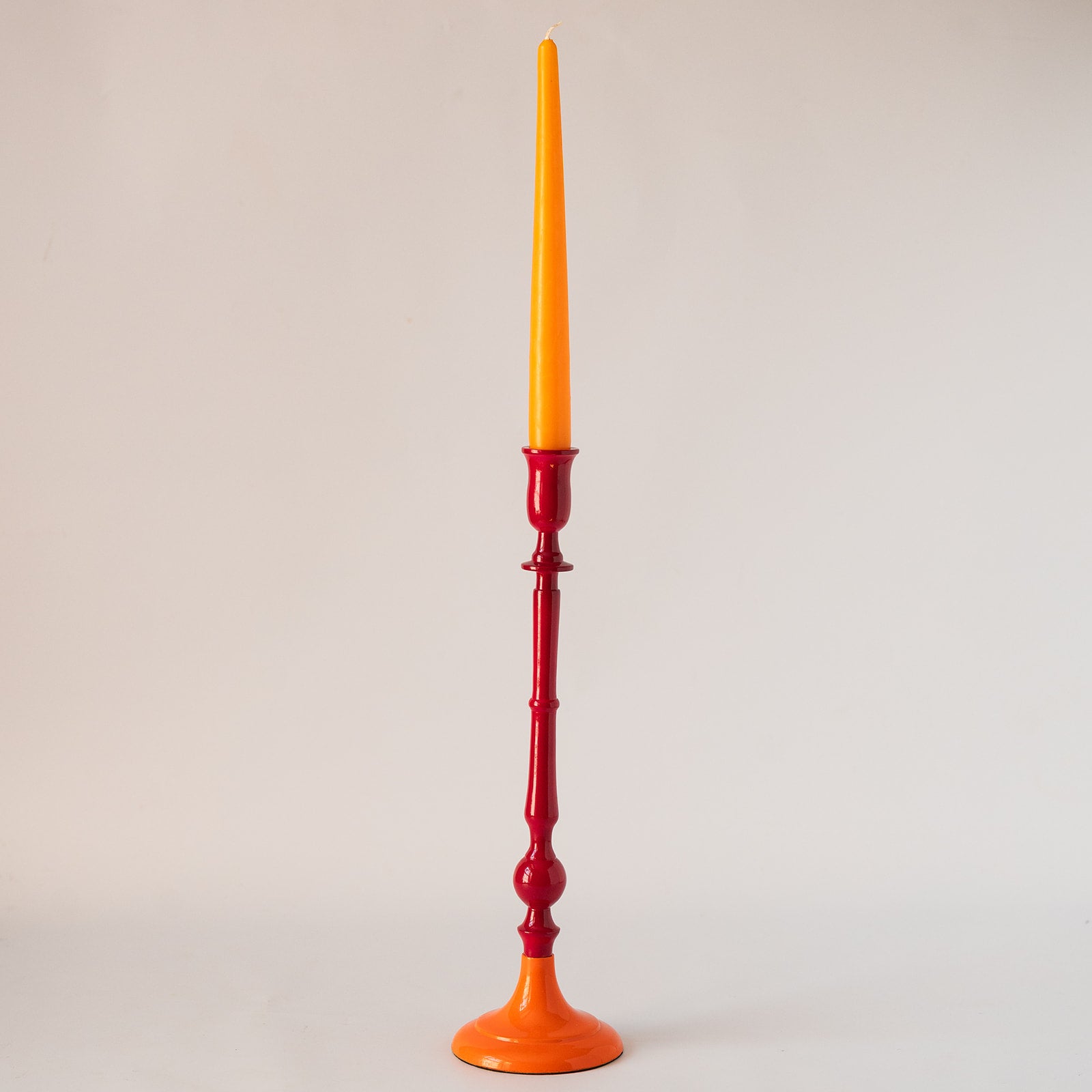 A tall candlestick holder with a glossy red stem and base, holding a long orange taper candle, photographed against a plain light gray studio background.