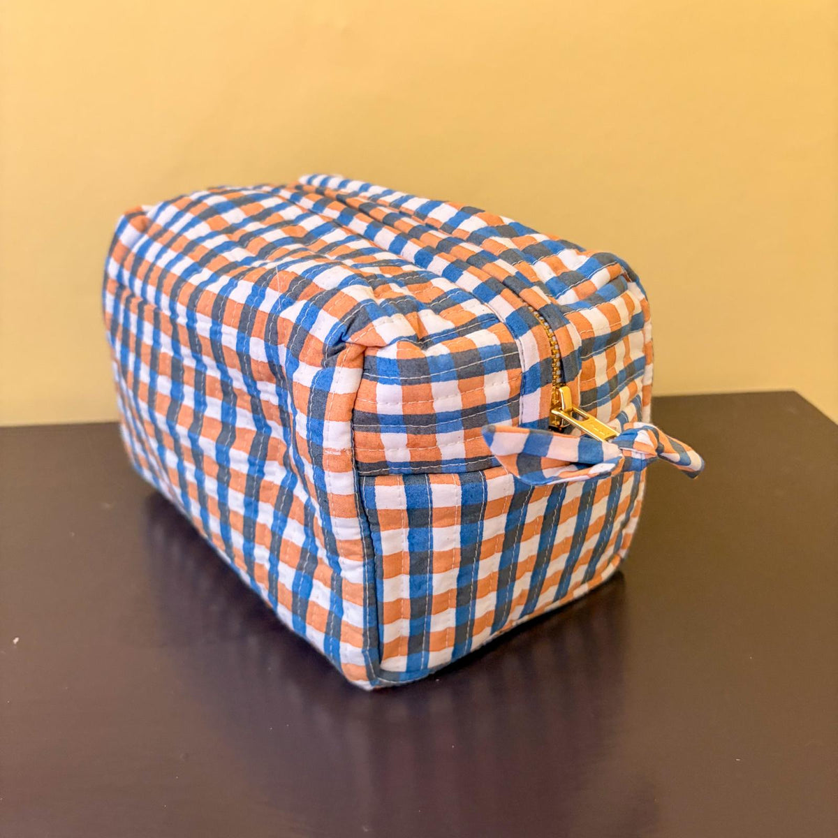 A square zippered pouch with a blue, orange, and white gingham checkered pattern, resting on a dark brown surface against a yellow background.