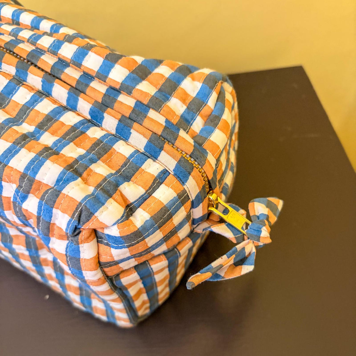 A close-up of a quilted fabric zippered pouch with a blue, orange, and white plaid pattern, featuring a matching fabric pull tab with a gold zipper pull, resting on a dark surface.