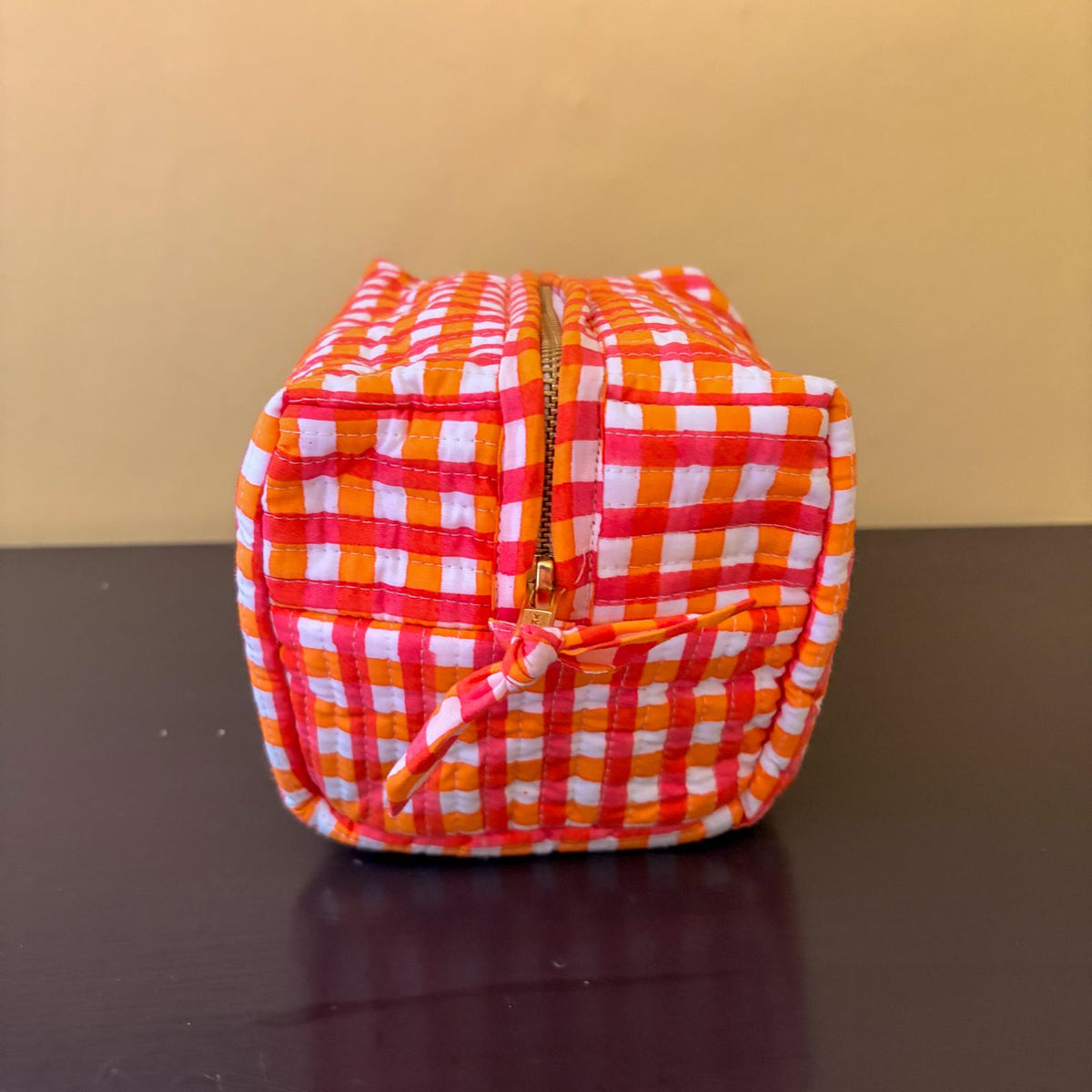 A small zippered cosmetic pouch with a bright orange and white gingham check pattern, resting on a dark brown surface against a beige background.