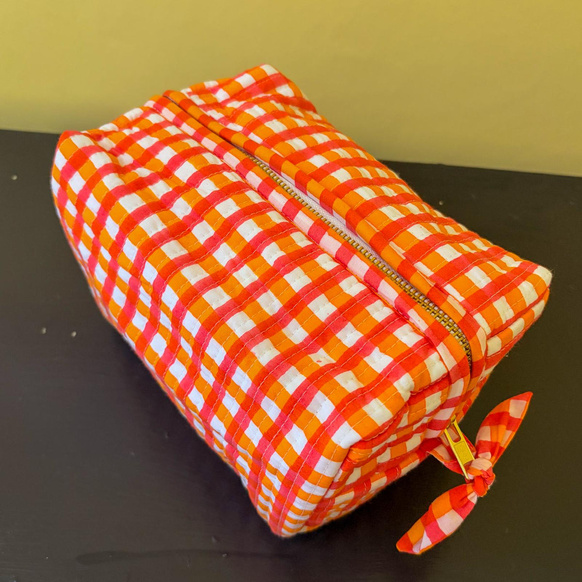 A rectangular zippered pouch with a vibrant orange, white, and red gingham pattern, resting on a dark surface against a yellow background.