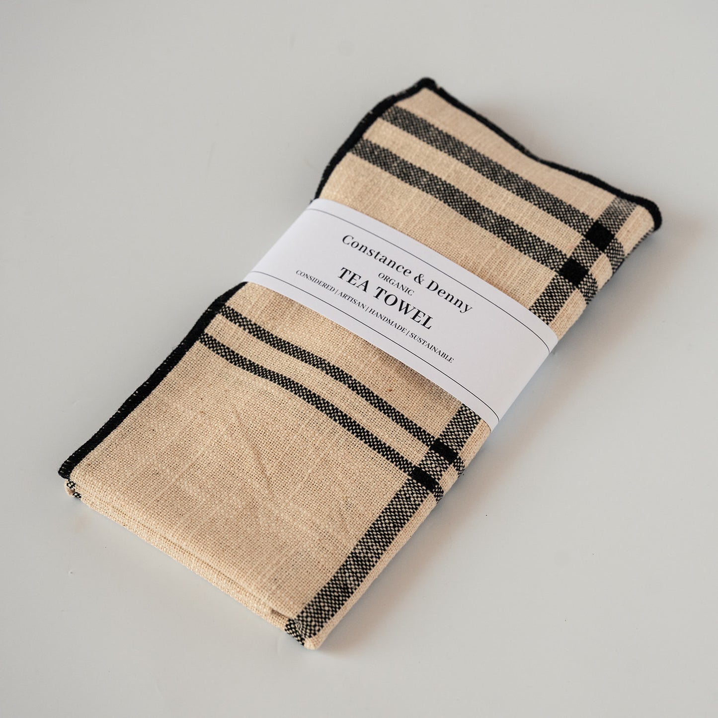 A folded Constance & Denny Organic Tea Towel in a beige and black plaid pattern, wrapped in a white paper band label, on a light gray background.