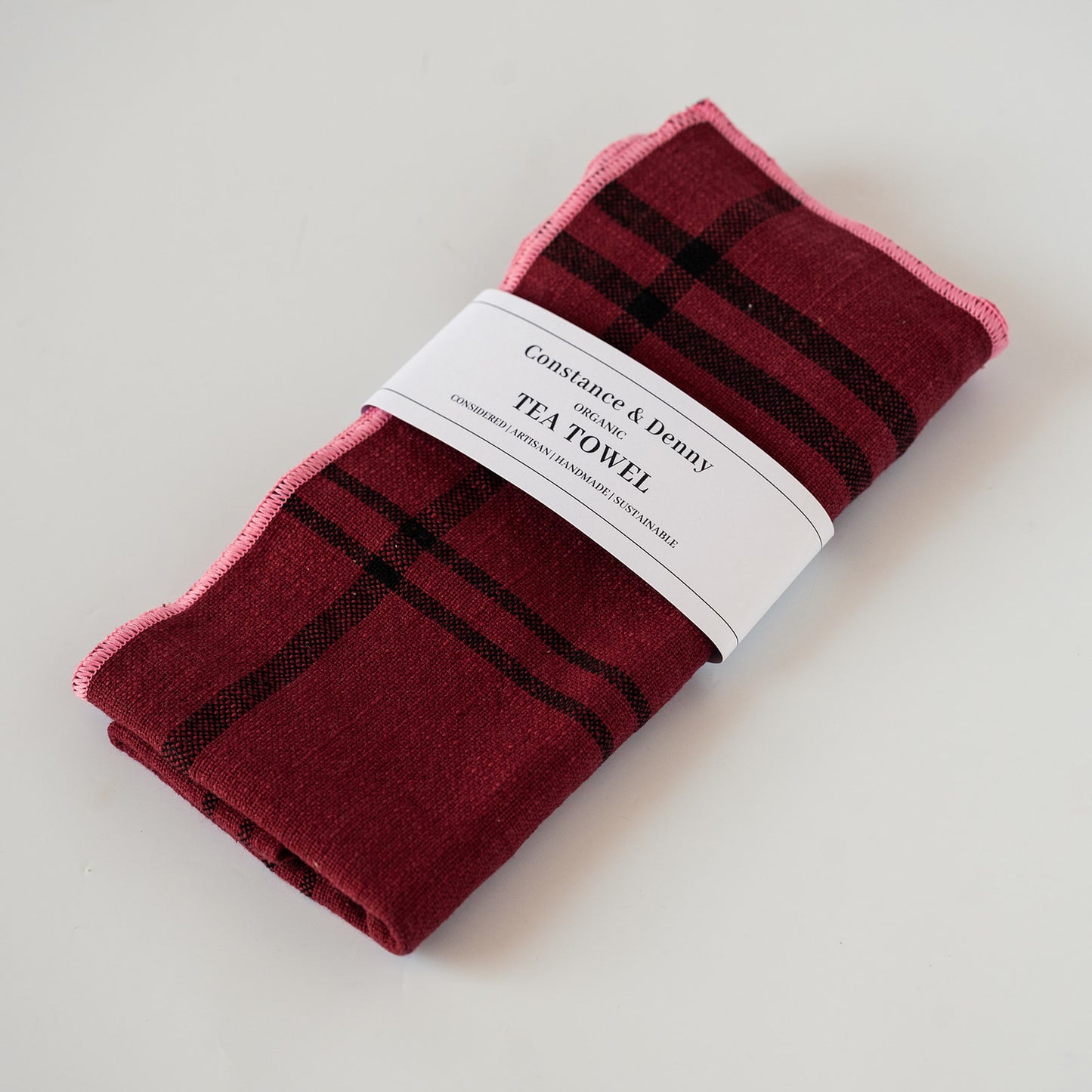 A folded tea towel with a dark red and black plaid pattern and a pink border, wrapped in a white paper band that reads 'Constance & Deny Tea Towel'.