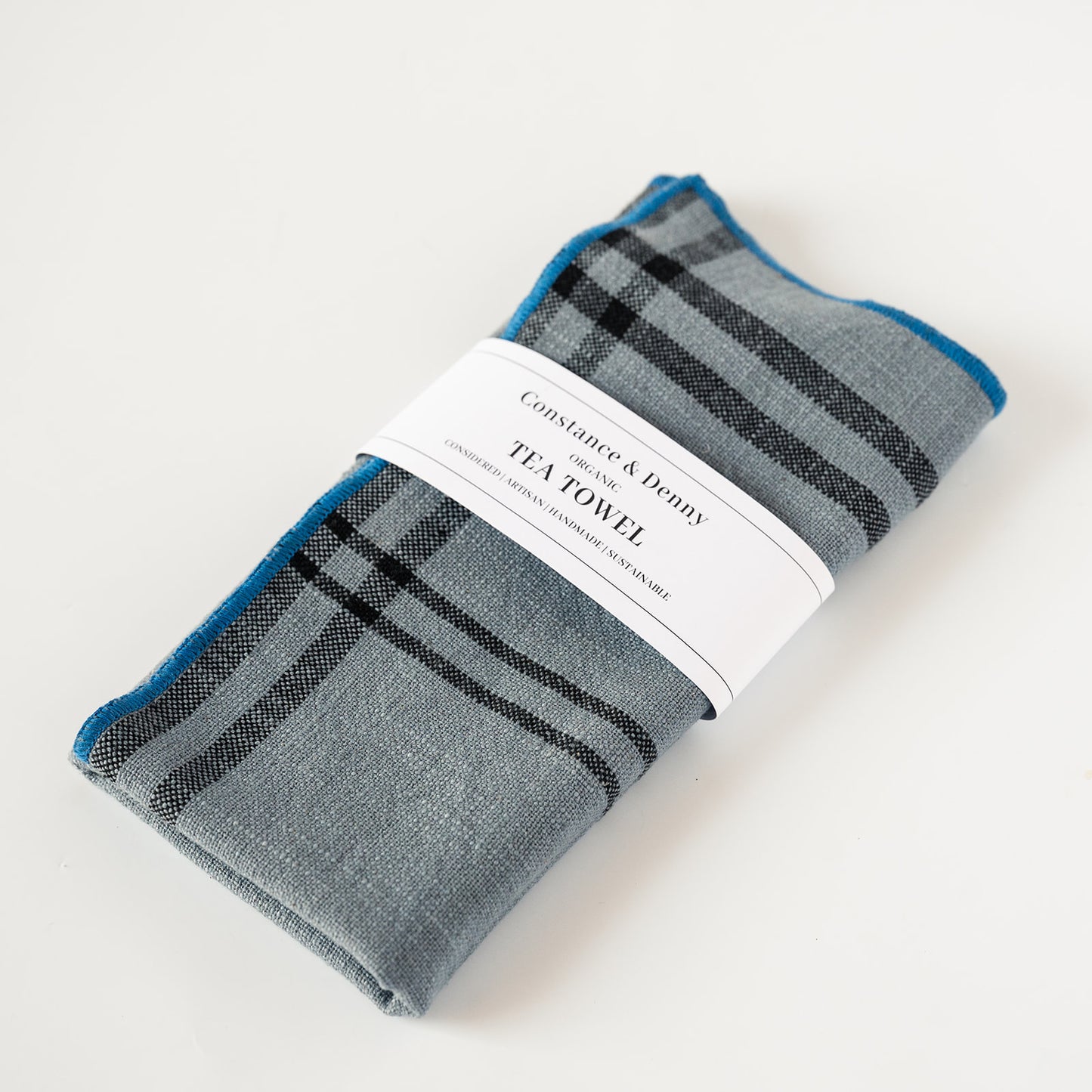 A folded gray tea towel with a black and blue plaid pattern, wrapped in a white paper band that reads 'Constantine & Deny TEA TOWEL'.