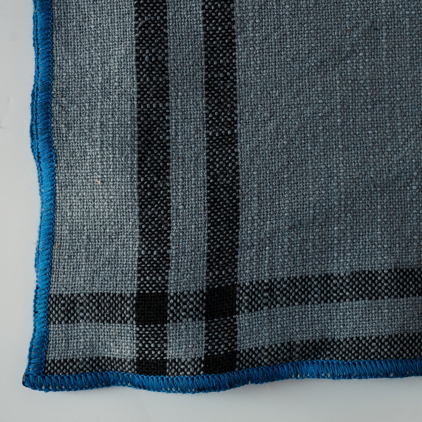 A close-up of a woven textile, likely a blanket, throw, or scarf, featuring a gray and black plaid pattern with a bright blue fringed edge.
