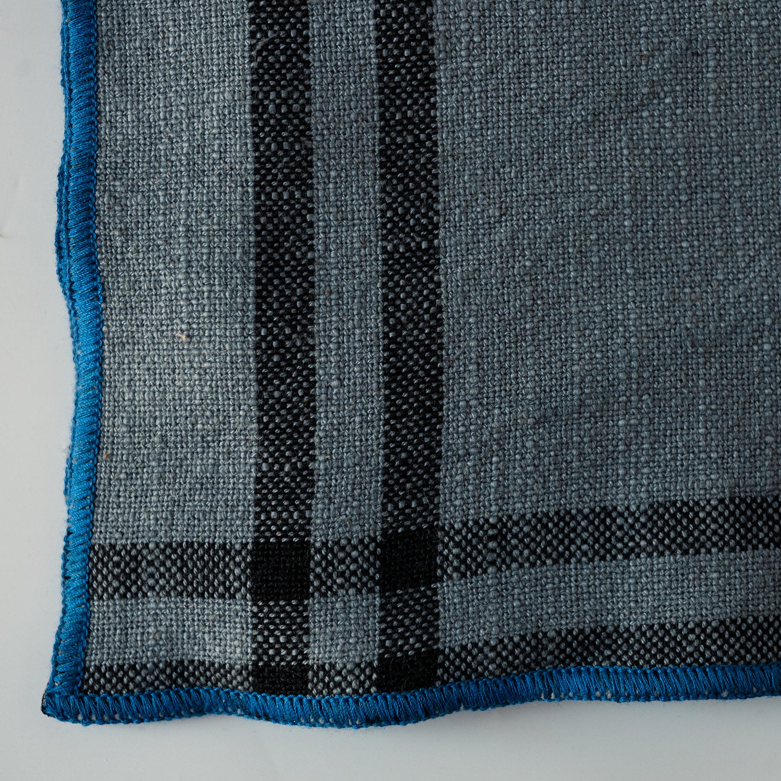 A close-up of a woven textile, likely a blanket, throw, or scarf, featuring a gray and black plaid pattern with a bright blue fringed edge.