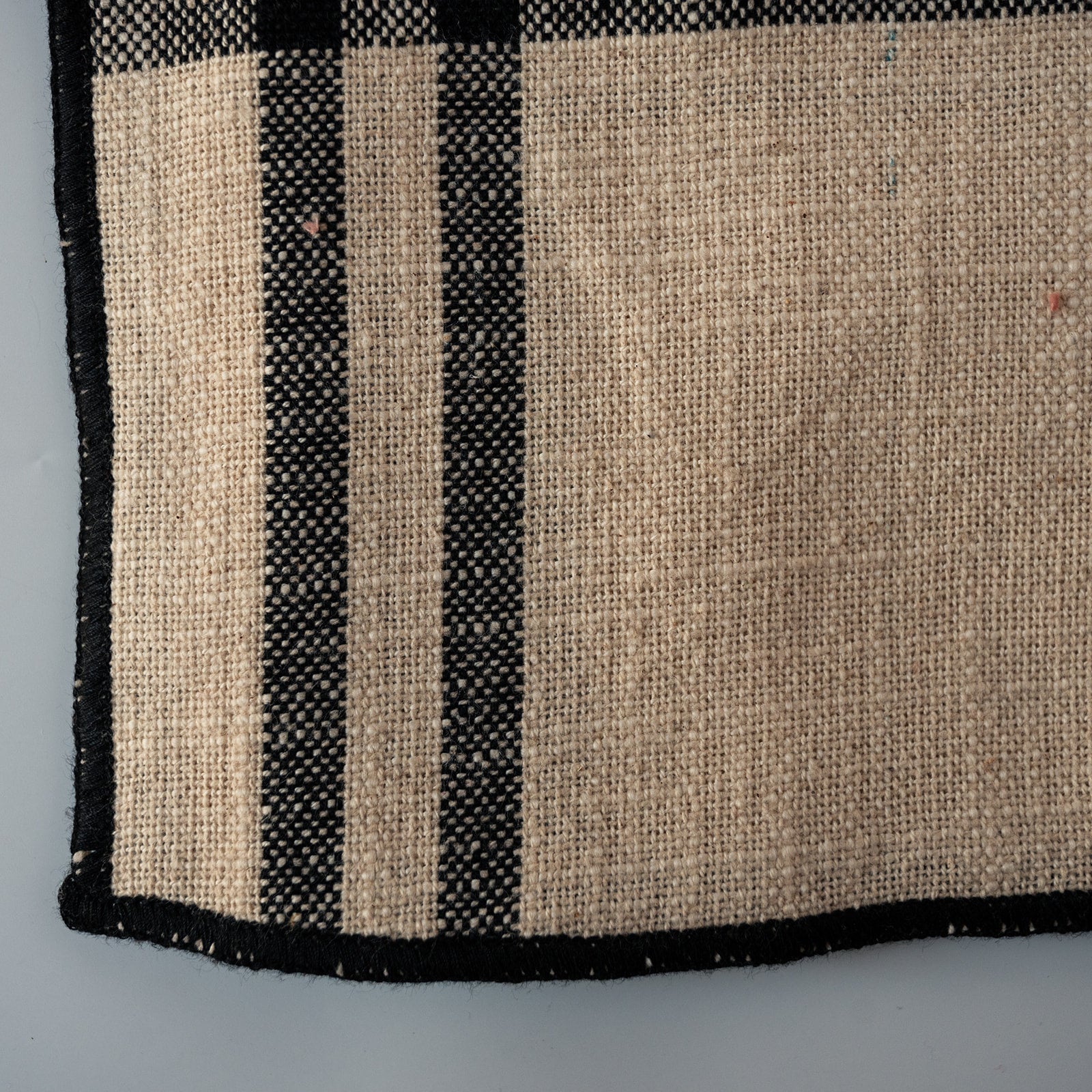 A close-up of a woven textile or rug corner showing a natural tan burlap-like weave with bold black stripes and a black border on a light gray background.