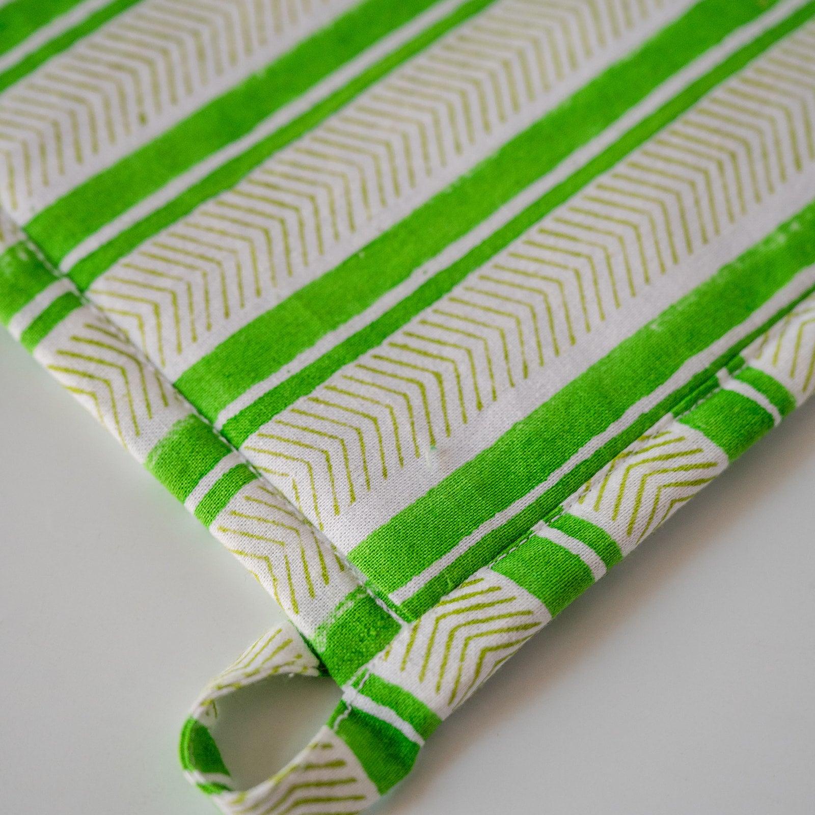 A close-up of a green and white striped fabric potholder with a stitched loop handle, showing a geometric chevron pattern.