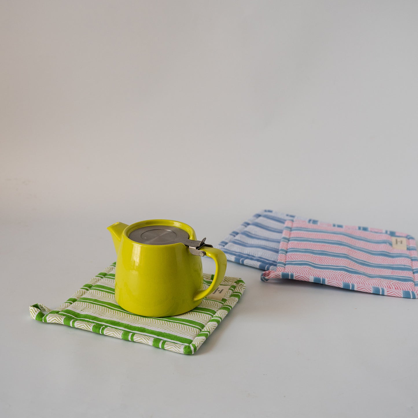 A bright yellow ceramic teapot with a metal infuser sits on a green and white striped cloth coaster. In the background, two folded striped kitchen towels or potholders, one blue and pink striped and one green and white striped, are stacked on a light gray surface against a white background.