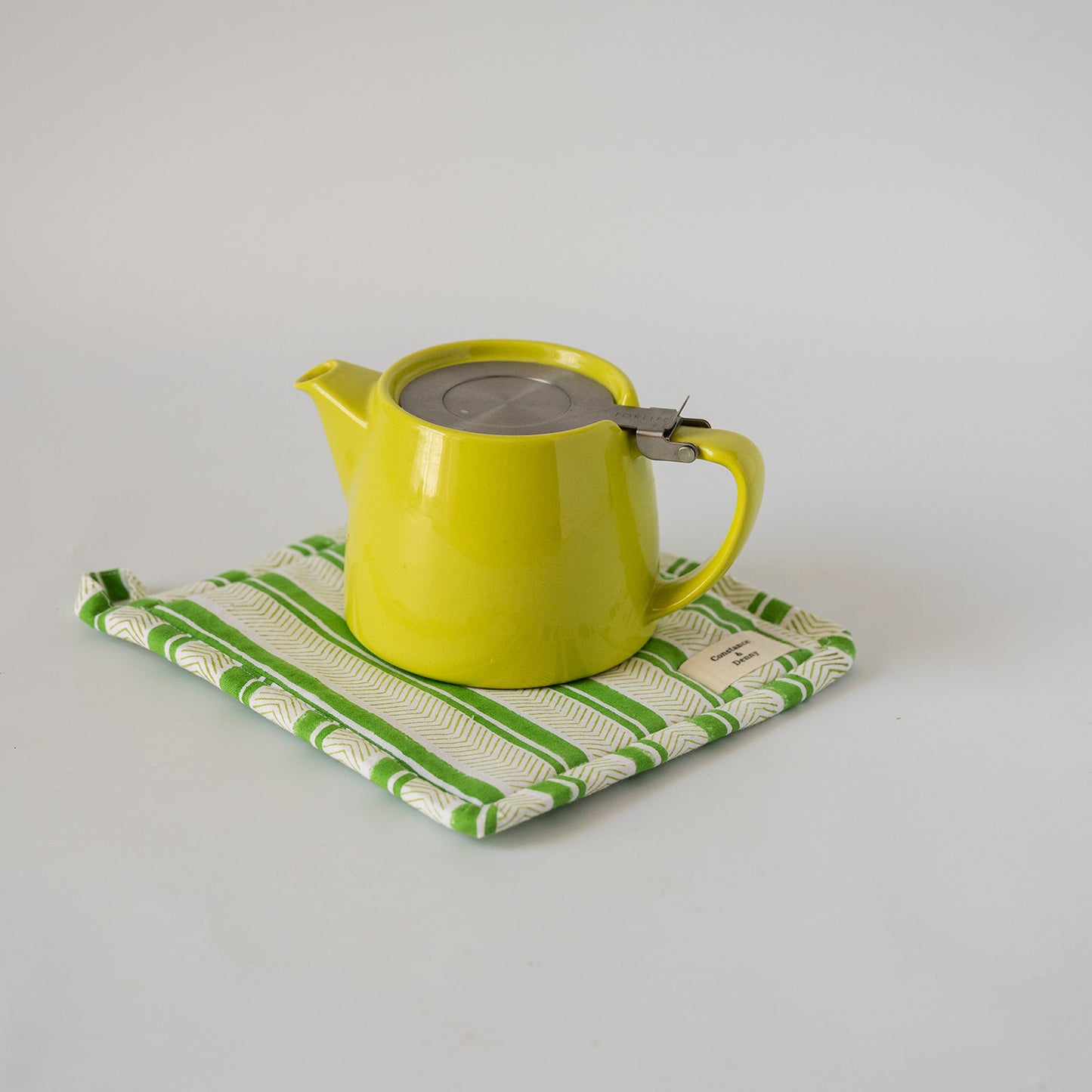 A bright yellow-green ceramic teapot with a stainless steel lid and metal infuser, resting on a green and white patterned cloth coaster against a plain light gray background.