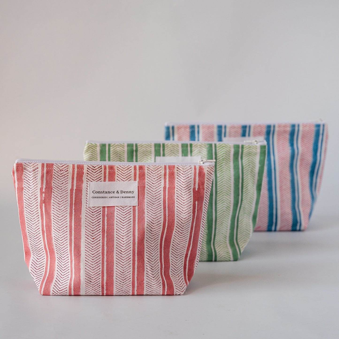 Three striped fabric pouches in coral, green, and blue tones arranged in a row on a light gray background. The front pouch has a white label reading 'Constance & Dreamy'.